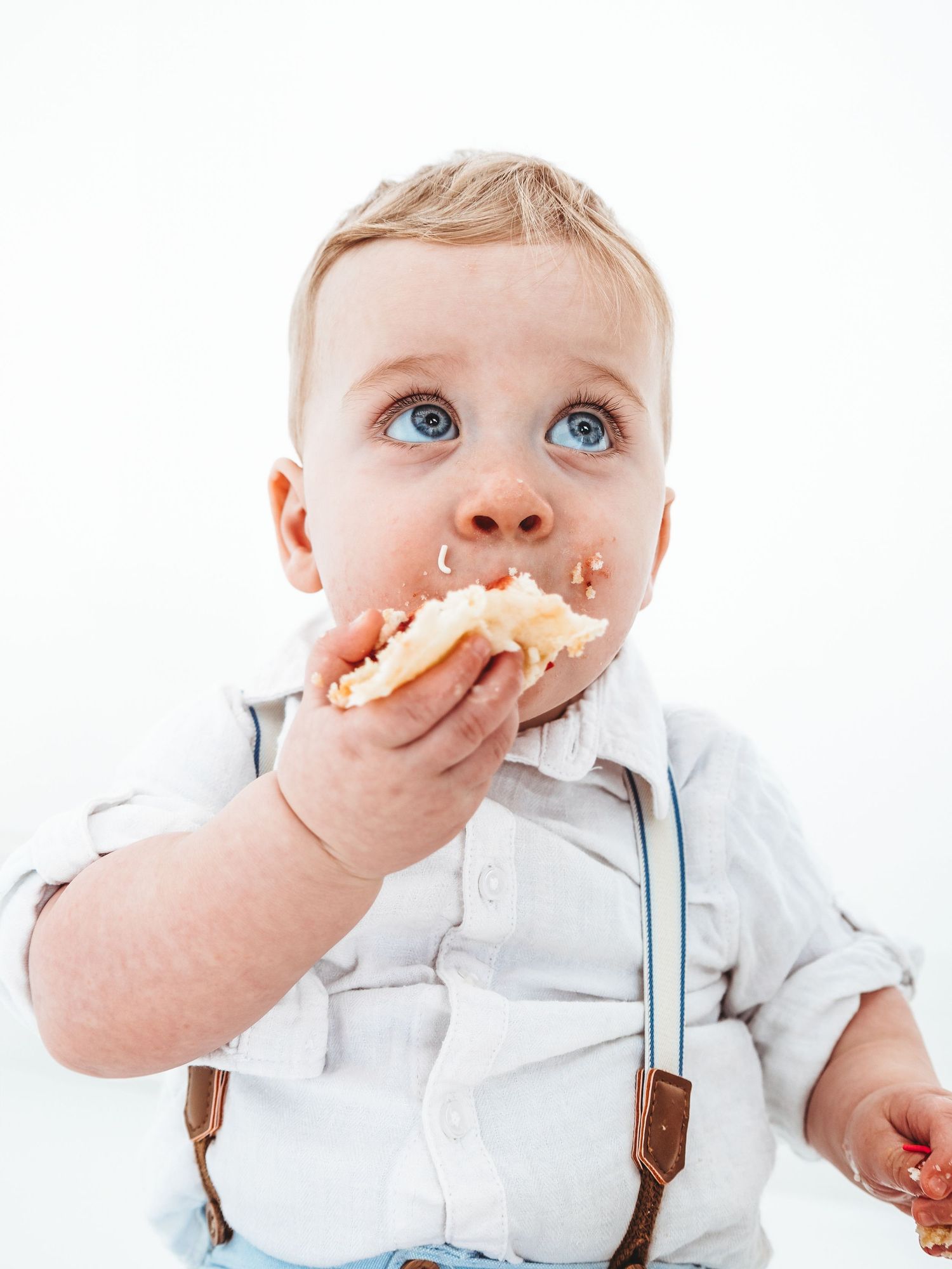 Cute blue-eyed toddler boy in suspenders eating cake with messy face on white background.