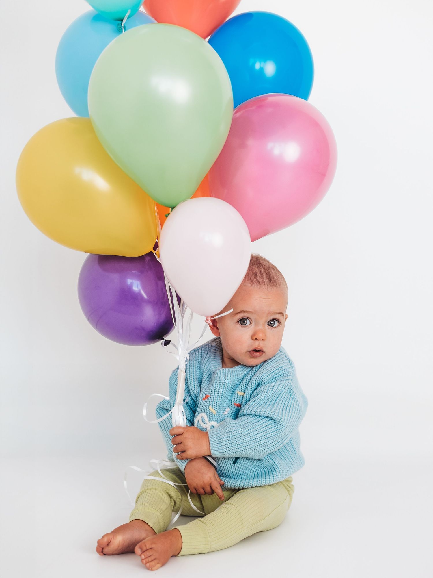 A cute baby in a blue sweater sits on white background holding a bunch of colorful balloons.