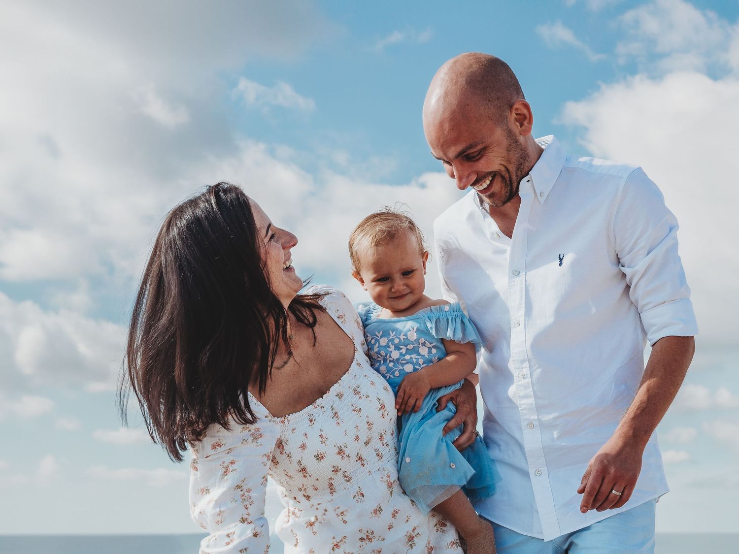 Happy family with mother, father, and baby smiling outdoors near the ocean under a partly cloudy sky.