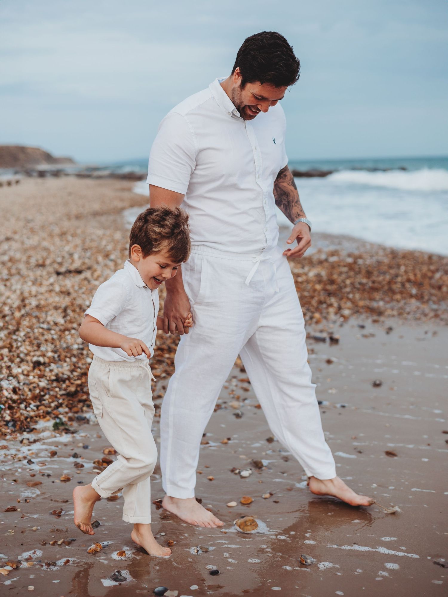 A father and young son walk barefoot on a pebble beach, both wearing matching white outfits, laughing together.