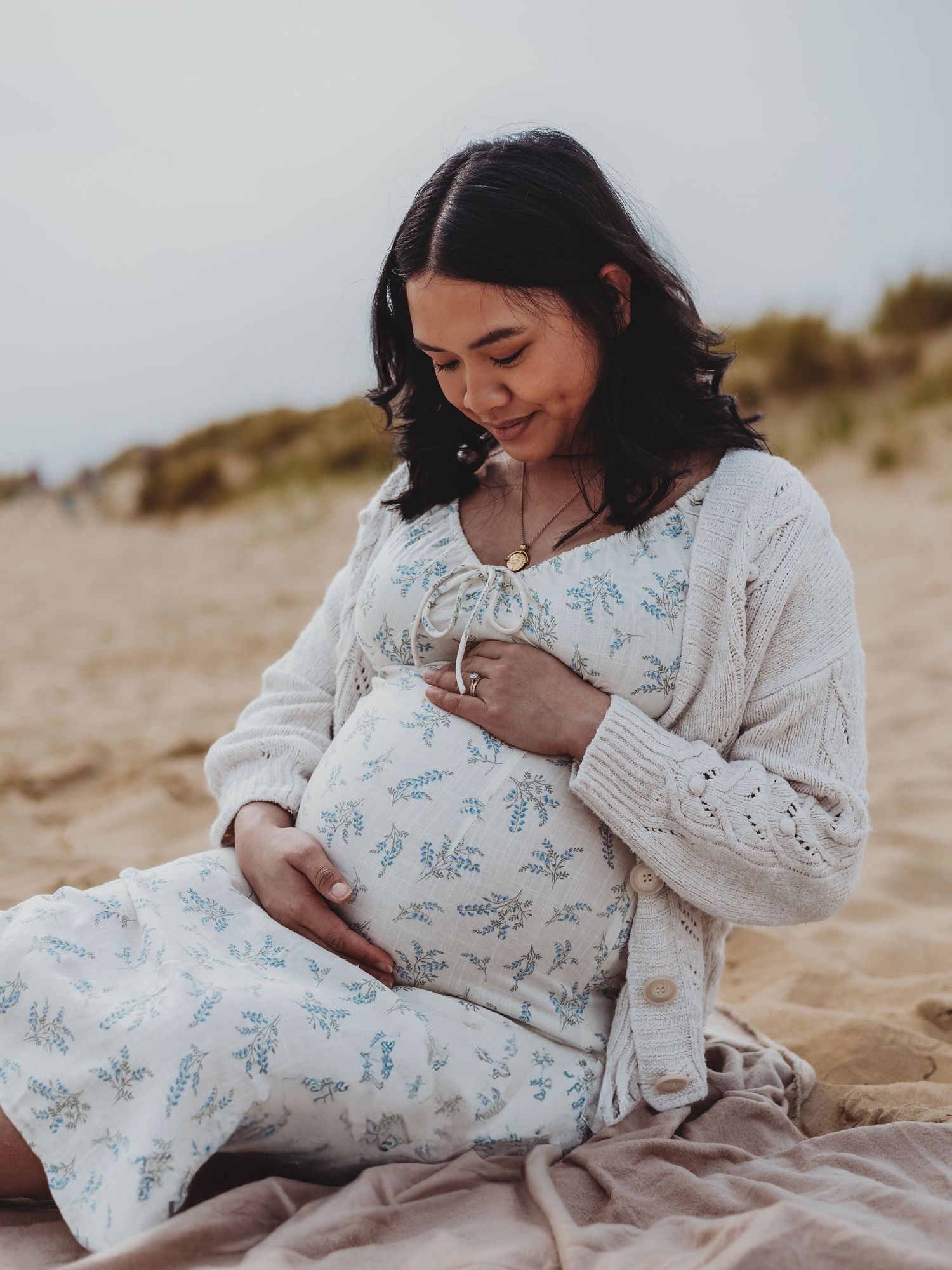 A mother in a white floral dress nurtures her newborn baby while sitting on a sandy beach blanket at sunset.