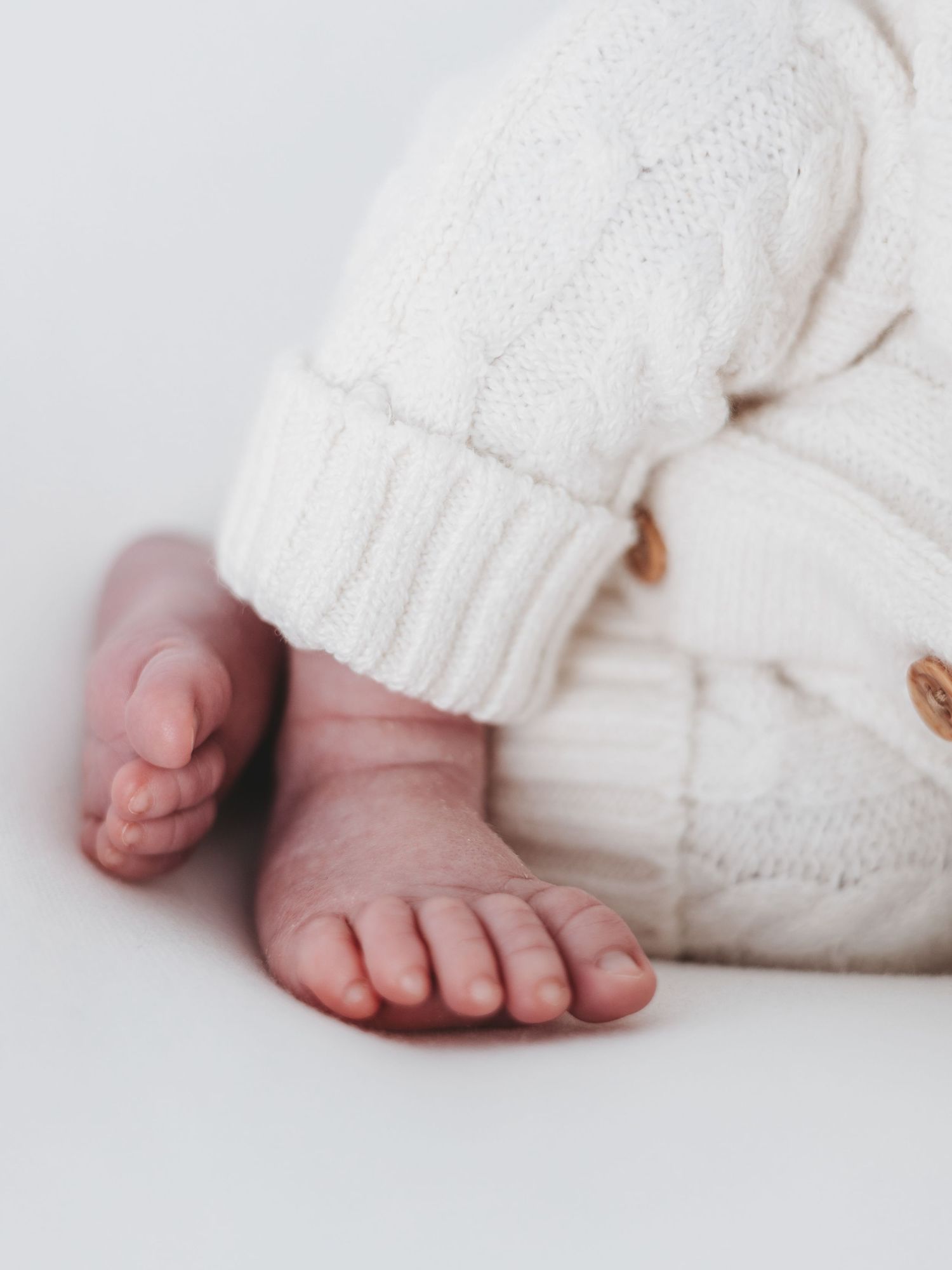 Tiny baby feet peek out from beneath a cozy white knit sweater with wooden buttons.