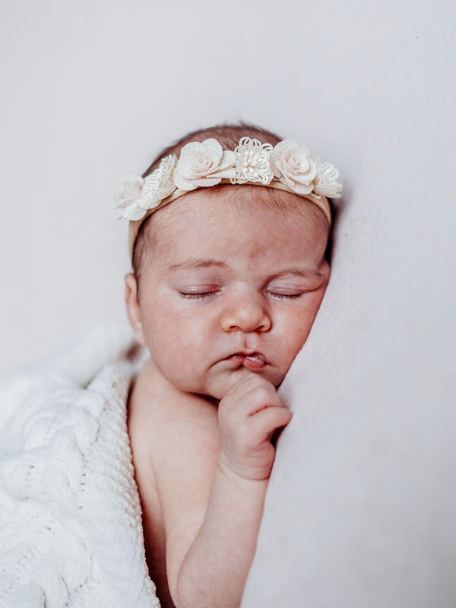 Sleeping newborn baby wearing white lace headband rests peacefully on white bedding.