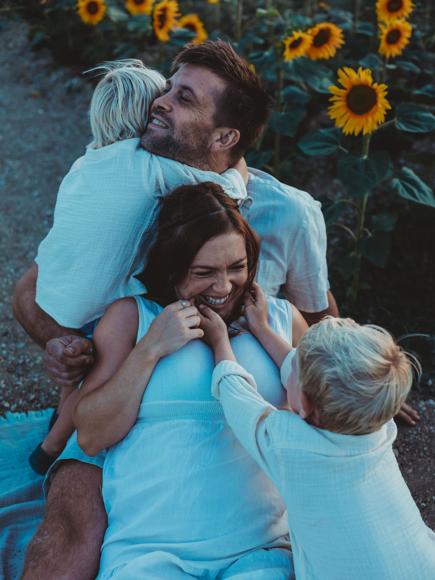 A loving family embraces in a sunflower field during golden hour, wearing coordinating light blue outfits.