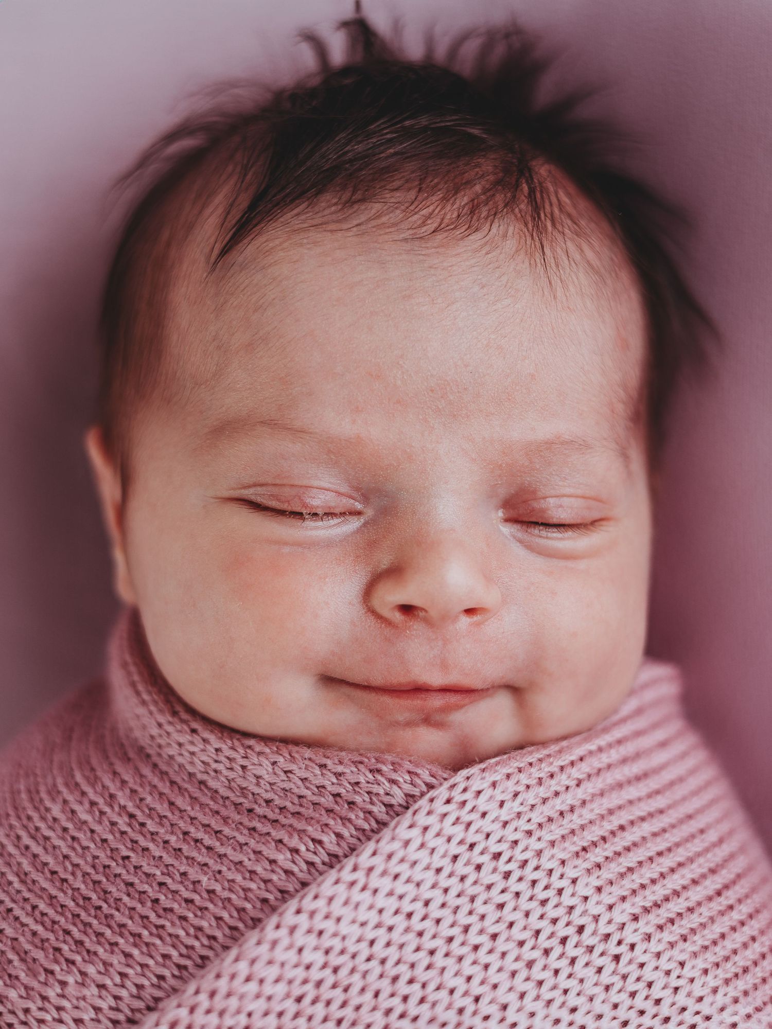 Peaceful sleeping newborn wrapped in soft pink knitted blanket with a gentle smile on their face.