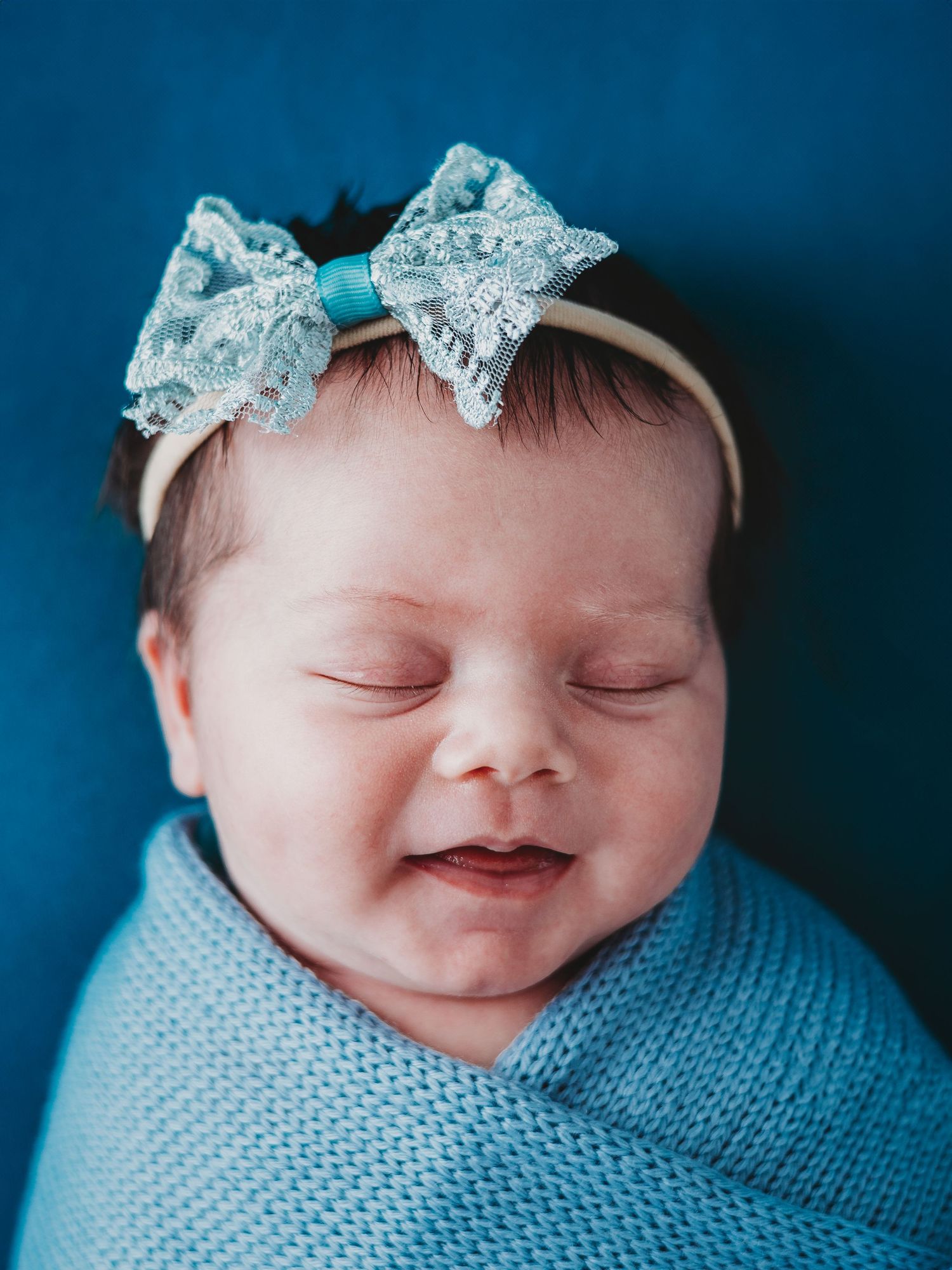 Sleeping baby wrapped in blue swaddle blanket wearing sparkly blue bow headband against navy background.