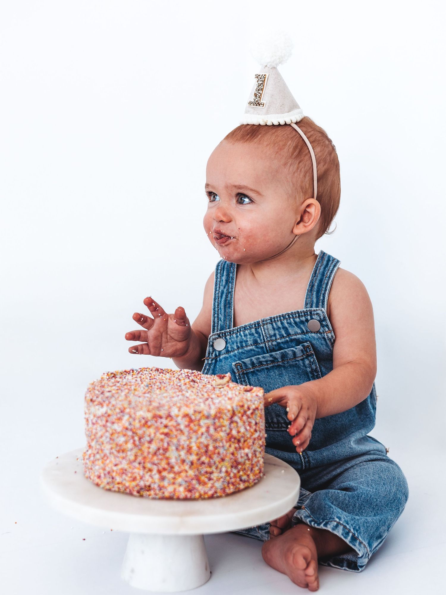 Baby in denim overalls wearing a party hat smashes a colorful sprinkle cake during a first birthday smash cake photo session.