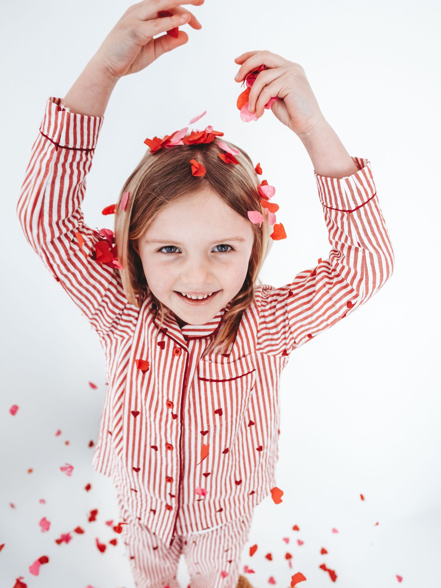 Young girl in red striped pajamas smiling joyfully while tossing red heart confetti in the air.