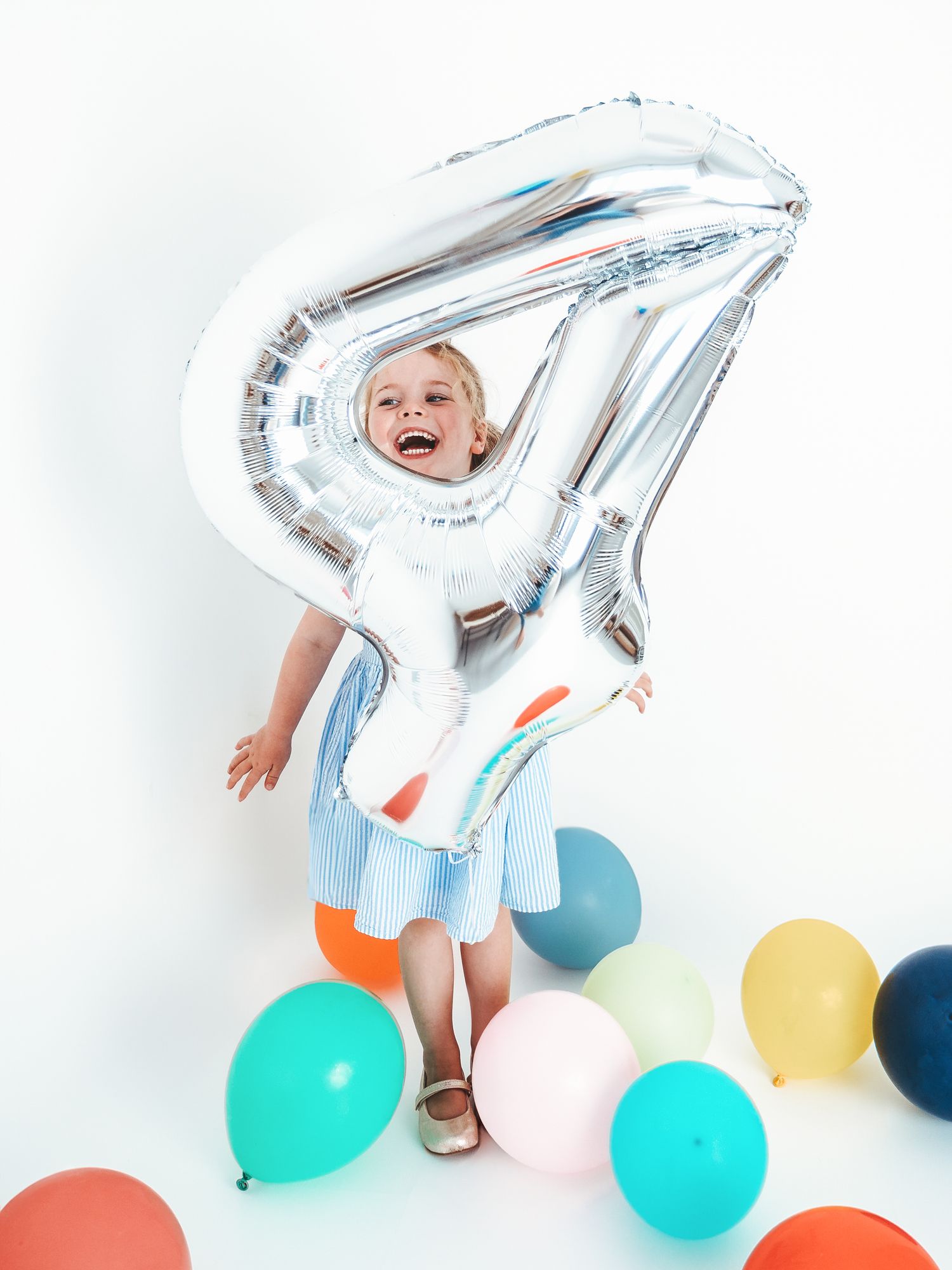 A laughing toddler holds a large silver number 4 balloon surrounded by colorful balloons at a birthday party.