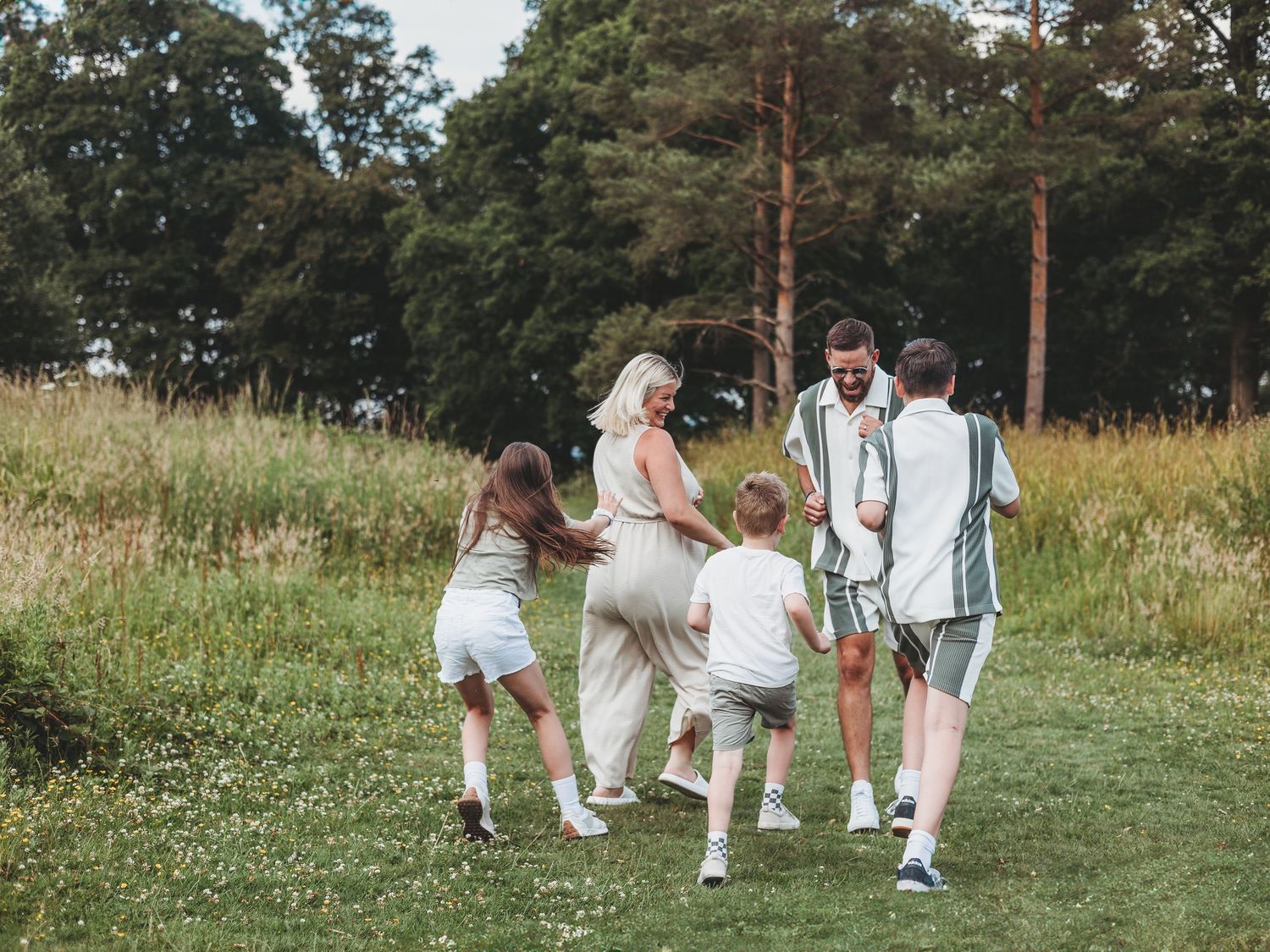 Family of five playing together on a lush green meadow surrounded by tall trees on a sunny day.