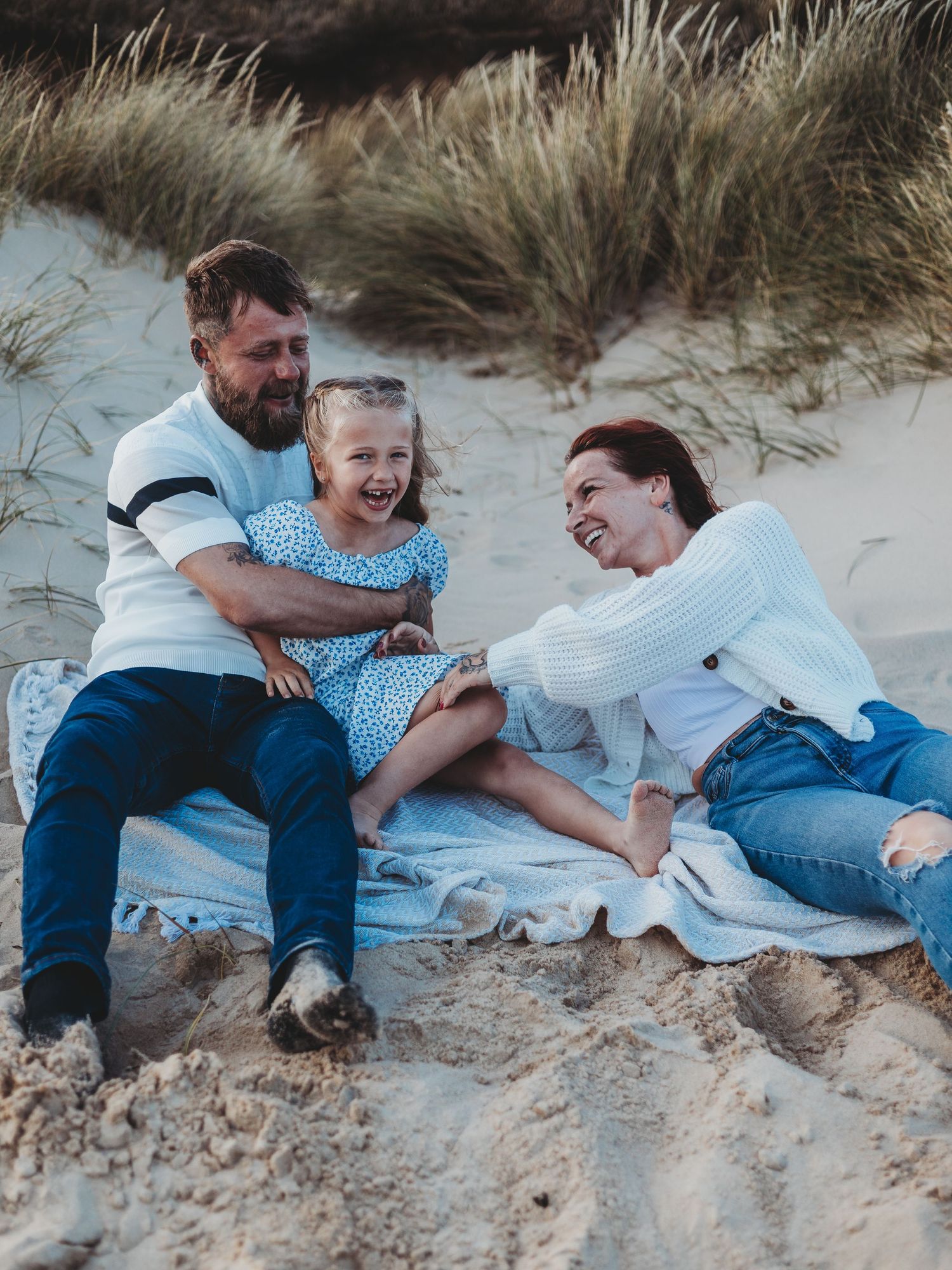 A joyful family of three laughing together on a sandy beach, with parents and young daughter sitting on a blanket near dunes.