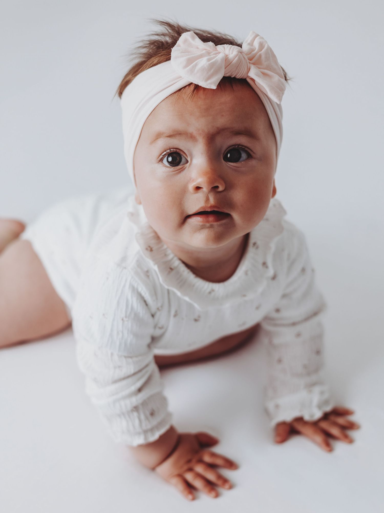 Adorable baby girl in white outfit and pink bow headband doing tummy time on white background.