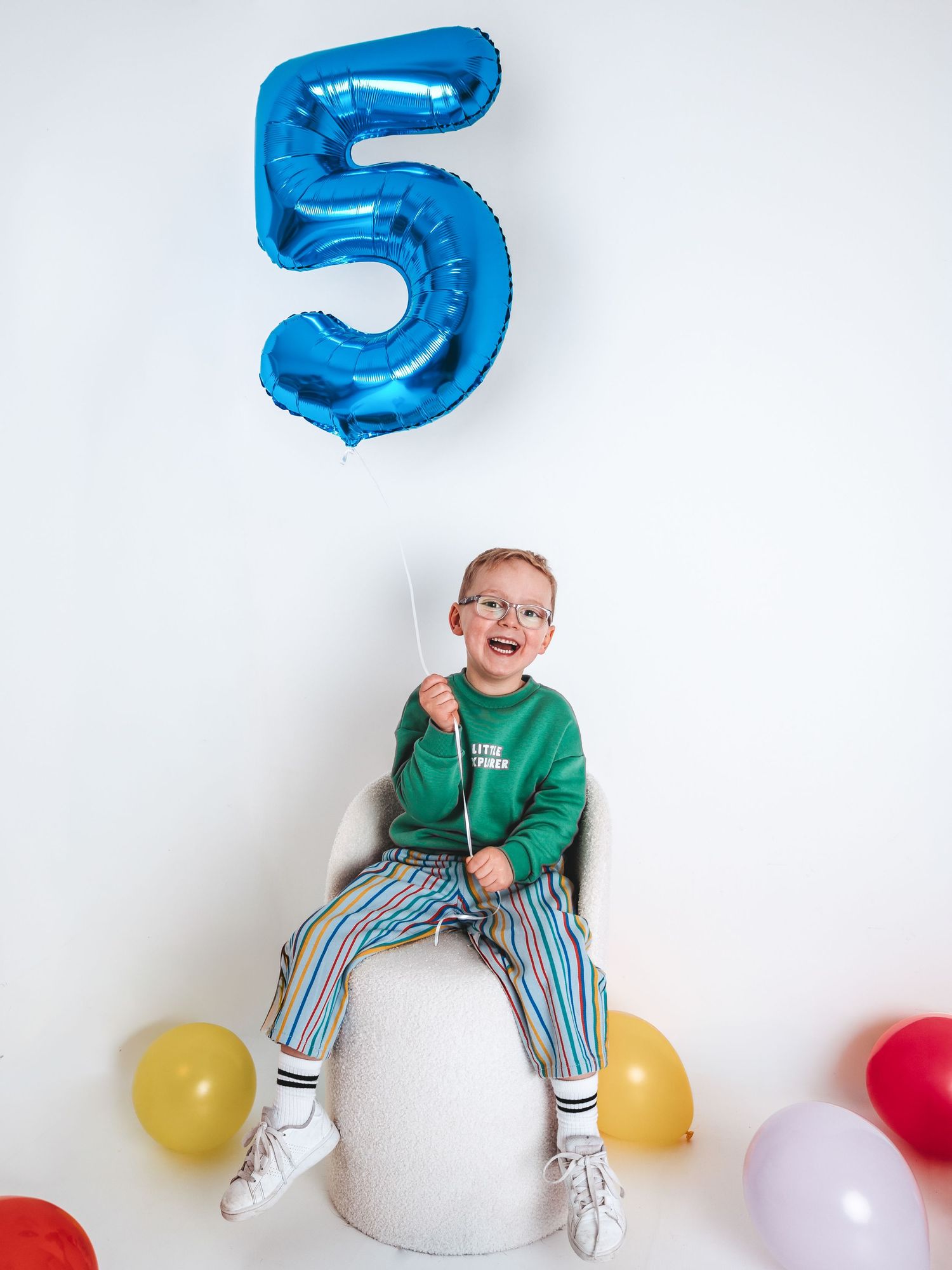 Happy boy celebrating 5th birthday, holding blue number 5 balloon, wearing green sweater and striped pants.