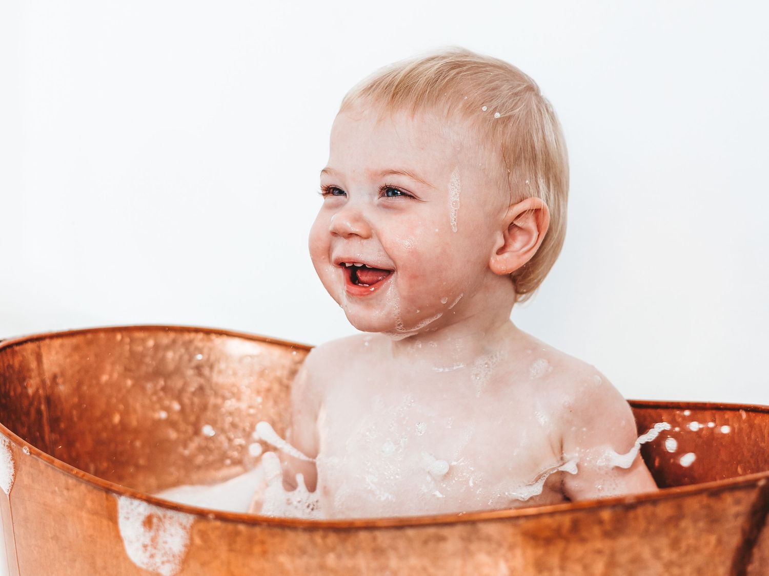 Happy baby laughing in a copper bathtub filled with soapy water against a white background.