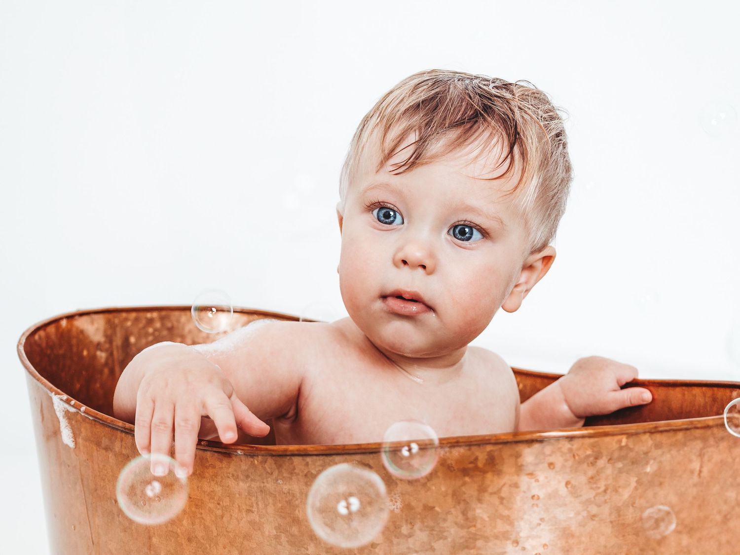 Cute baby with blue eyes sitting in a copper bathtub surrounded by soap bubbles on a white background.