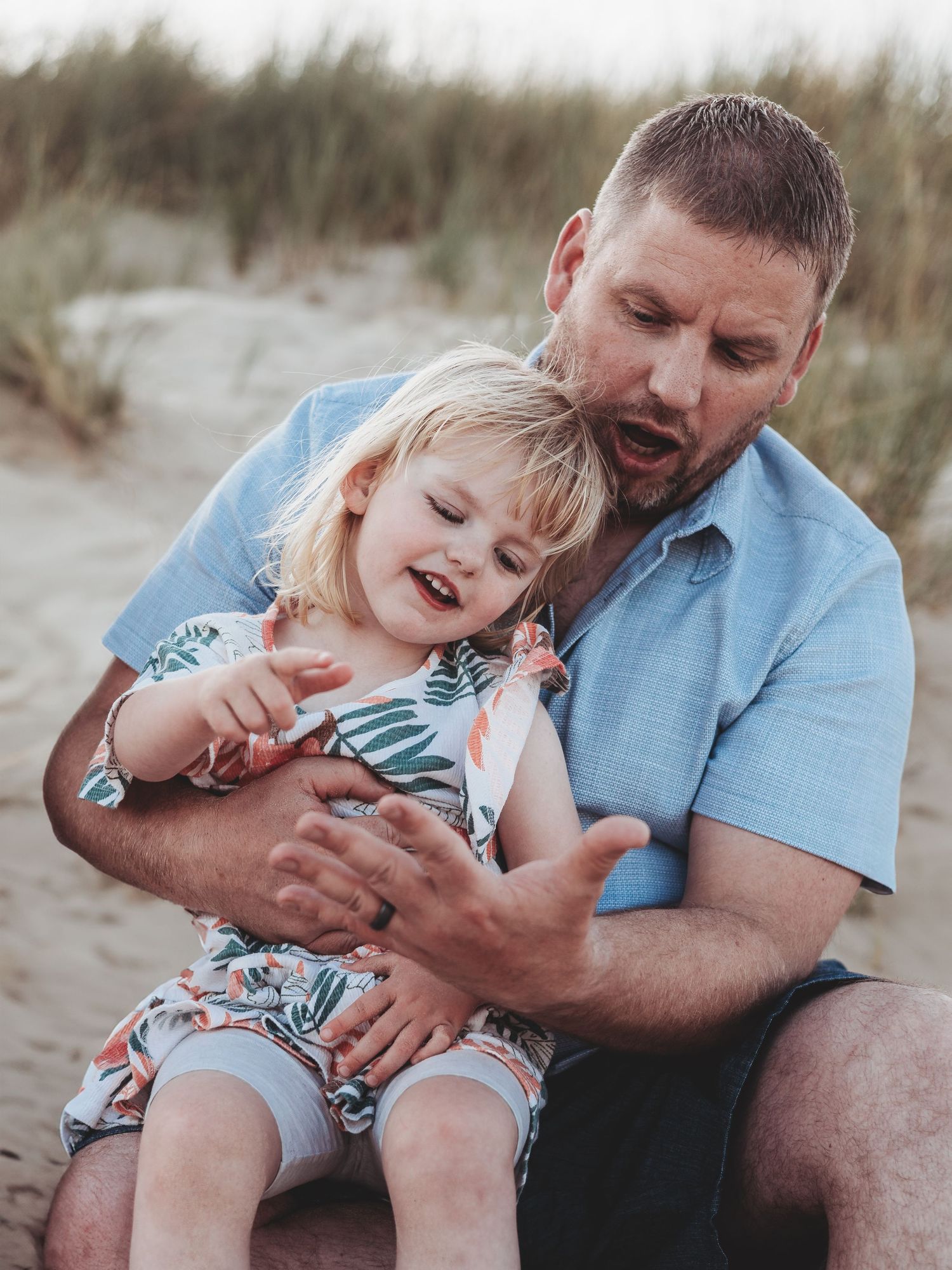 A father laughs and plays with his young blonde daughter on a sandy beach, both wearing casual summer clothing.
