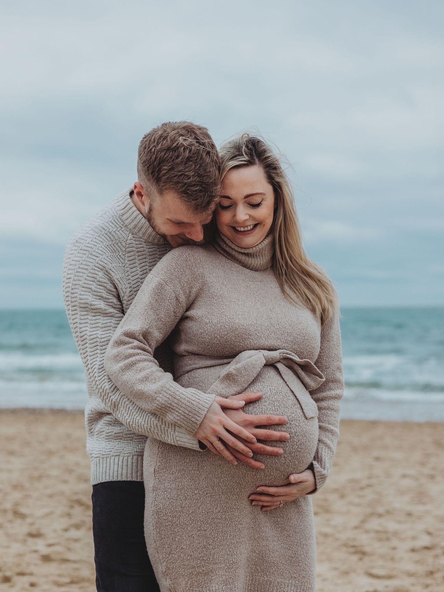 A couple in matching beige sweaters shares tender moments on a cloudy beach during a maternity photoshoot.