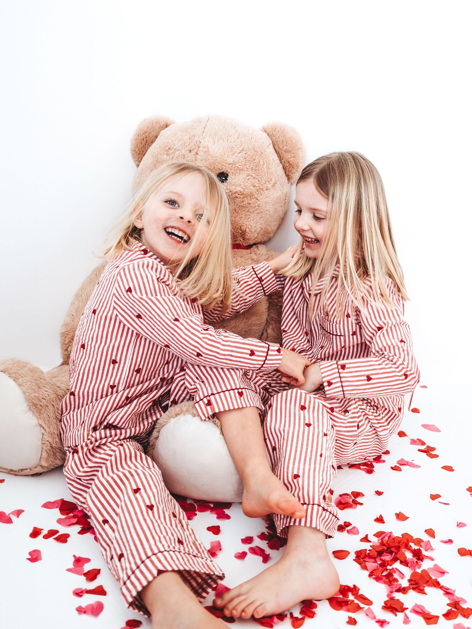 Two girls in matching red striped heart pajamas laugh together beside a giant teddy bear surrounded by rose petals.