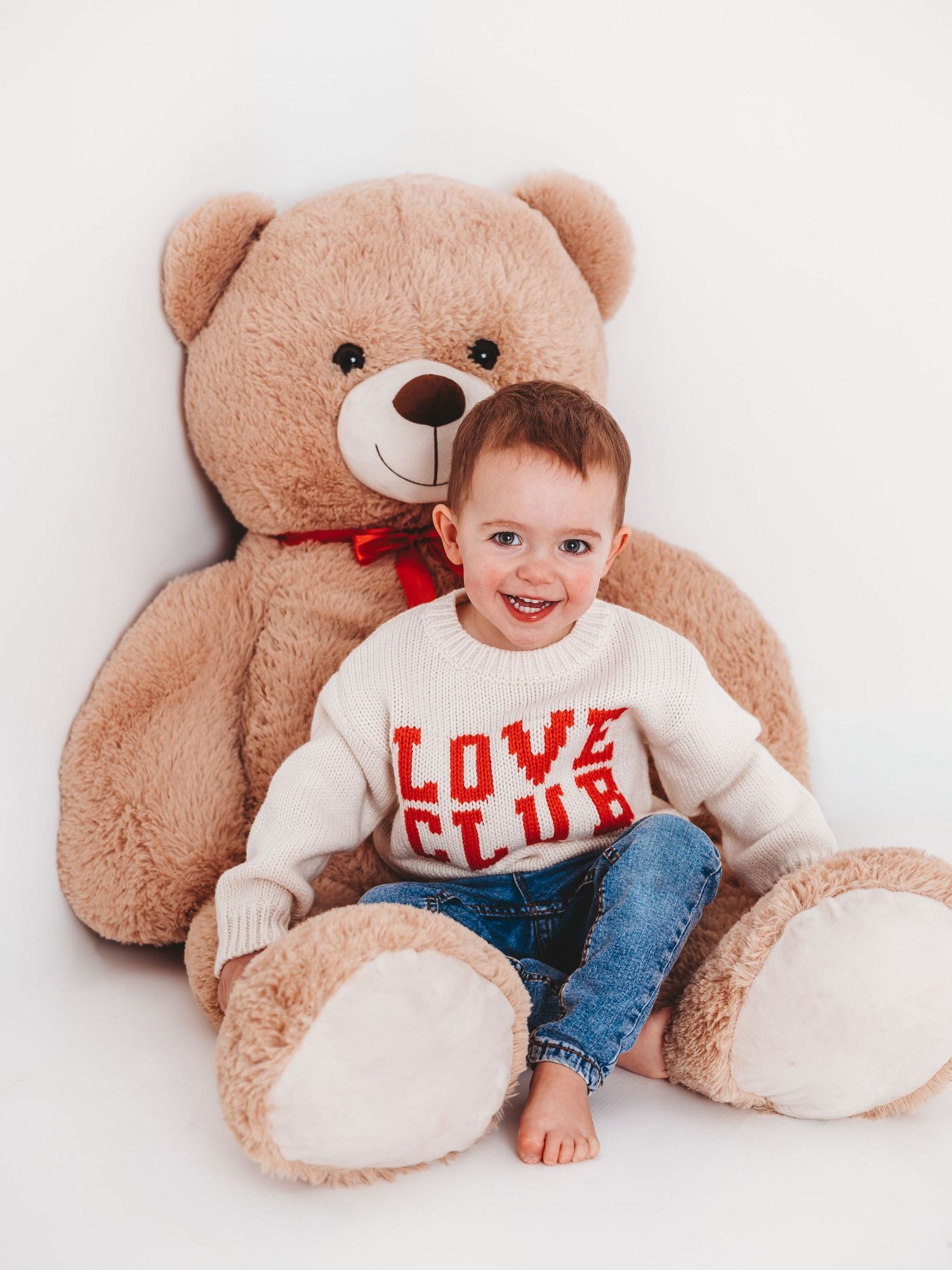 Smiling toddler in a white Love Club sweater and jeans sits in front of a giant teddy bear with a red bow.