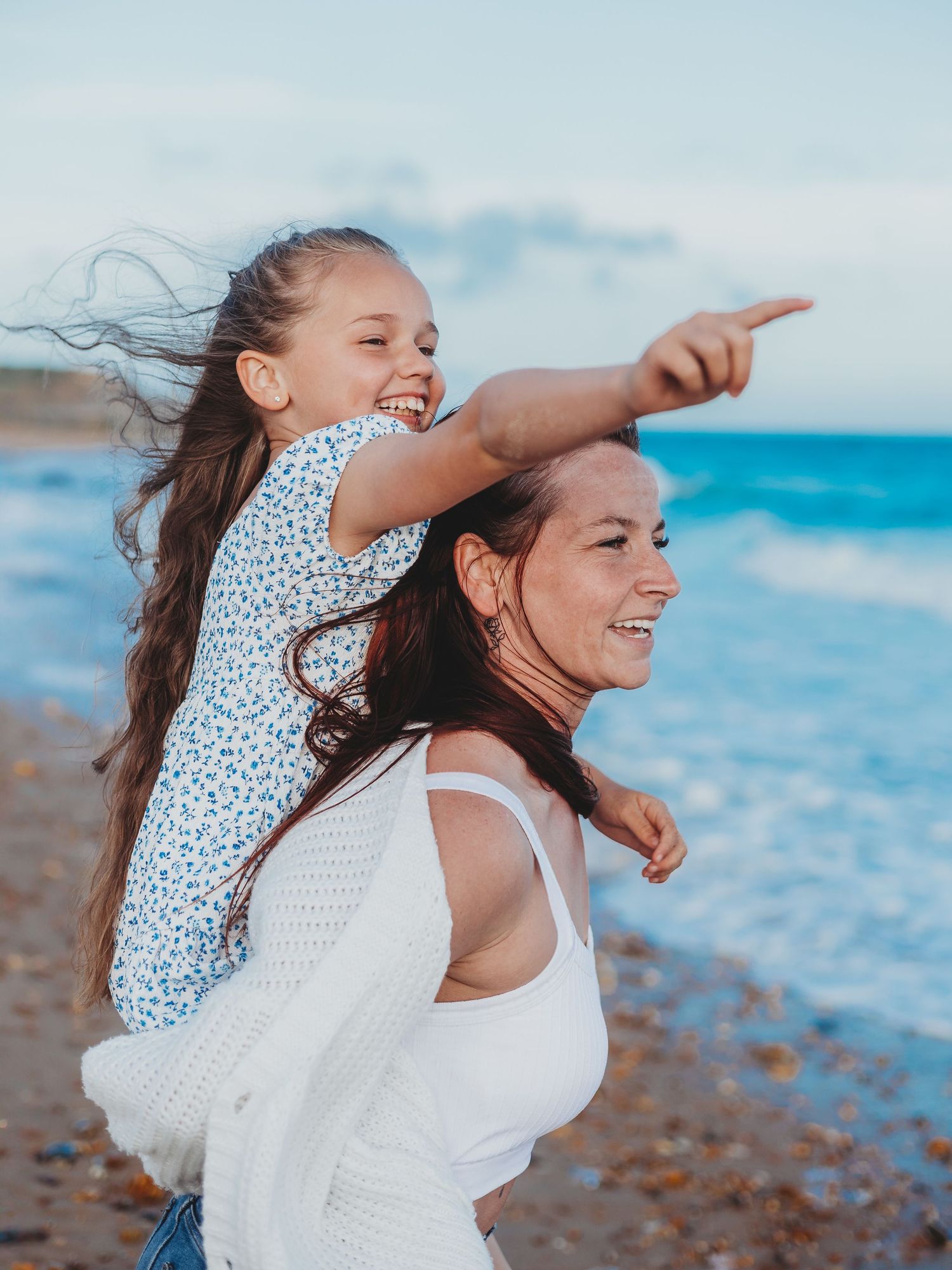 Happy girl sitting on mothers shoulders pointing at ocean on sunny beach, both smiling with wind in hair.