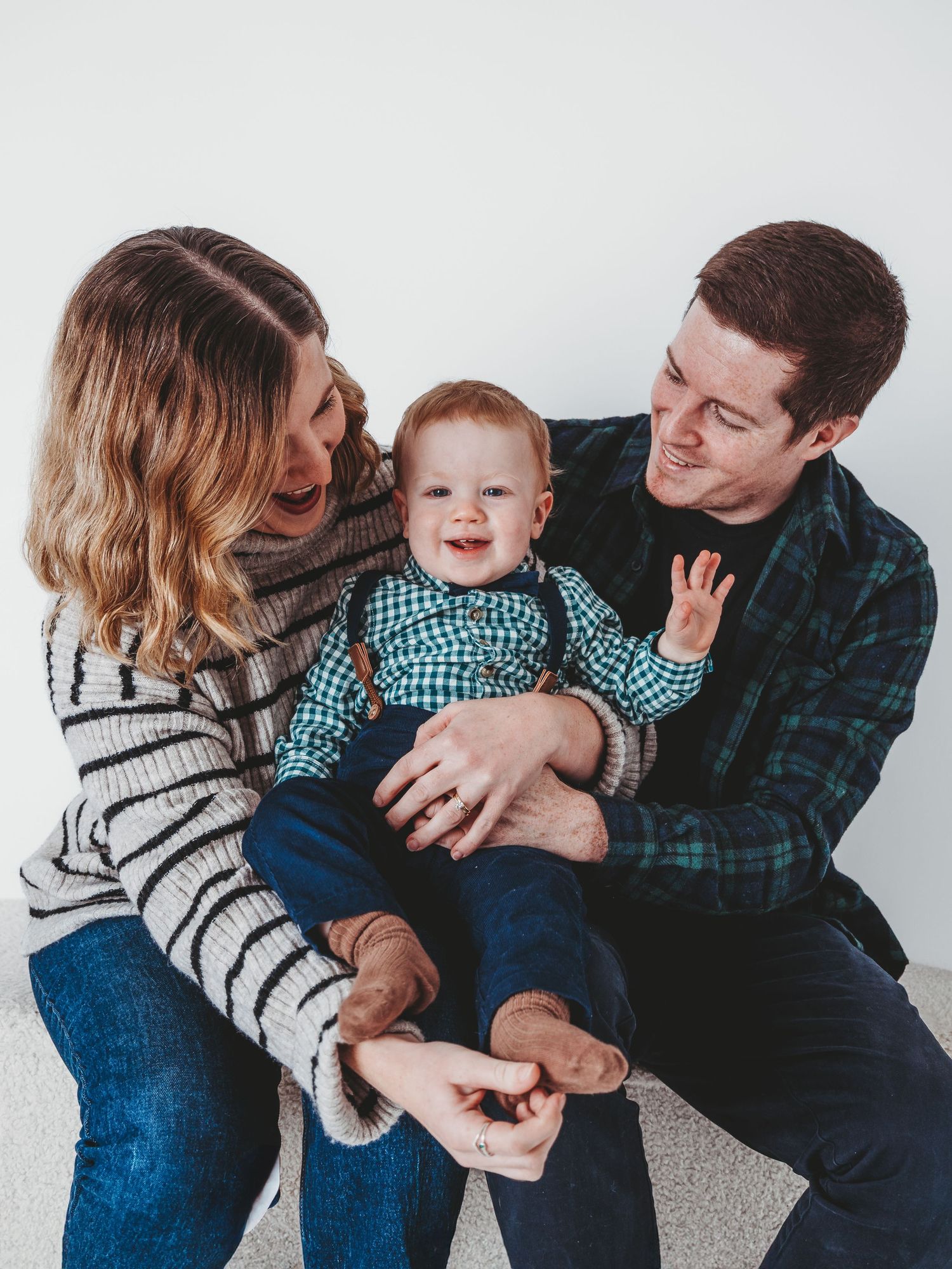 Happy family portrait with parents smiling at adorable red-haired baby boy in teal outfit against white background.