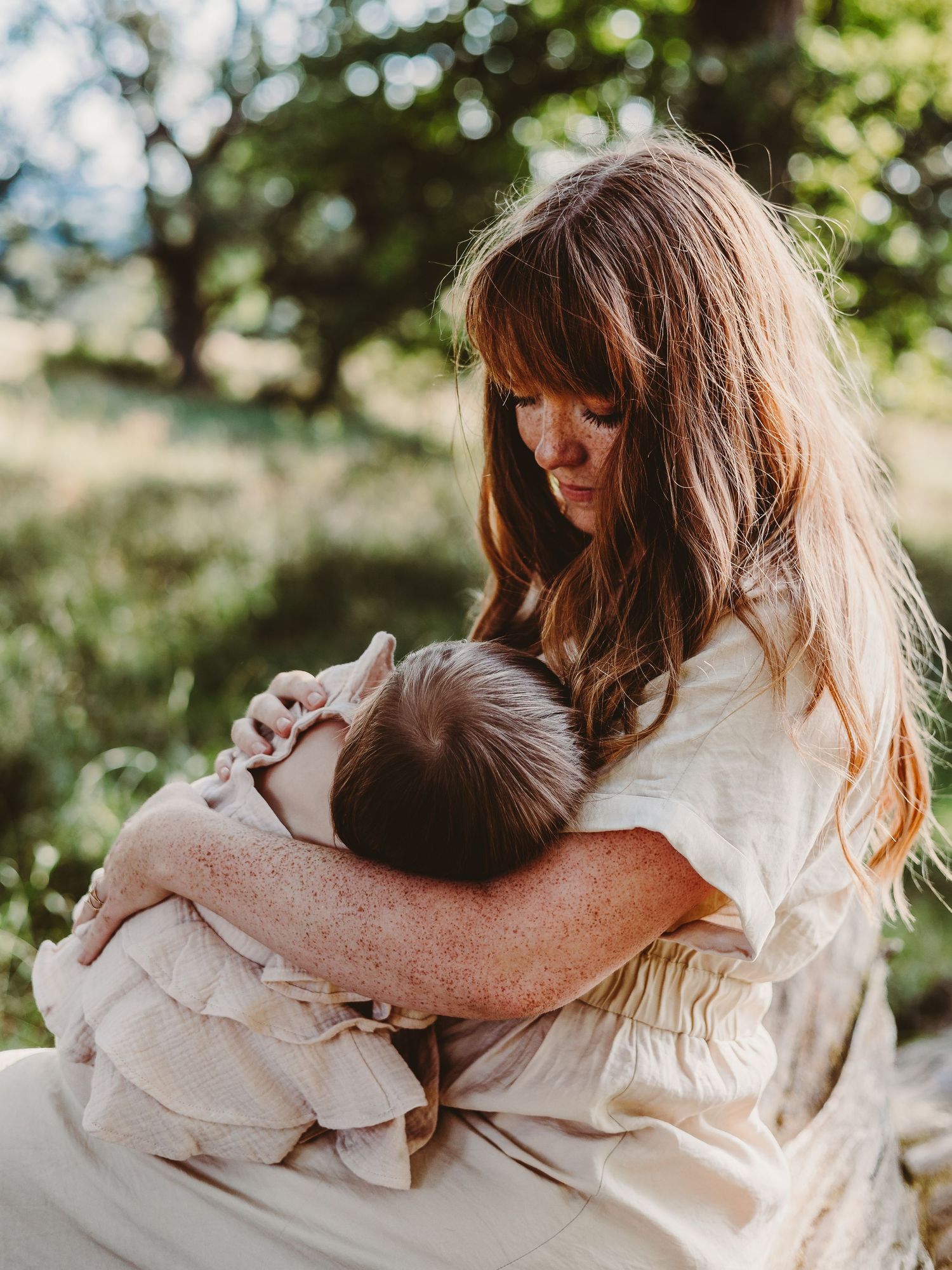 A mother with long auburn hair tenderly holds her newborn baby outdoors in a sunlit green garden.