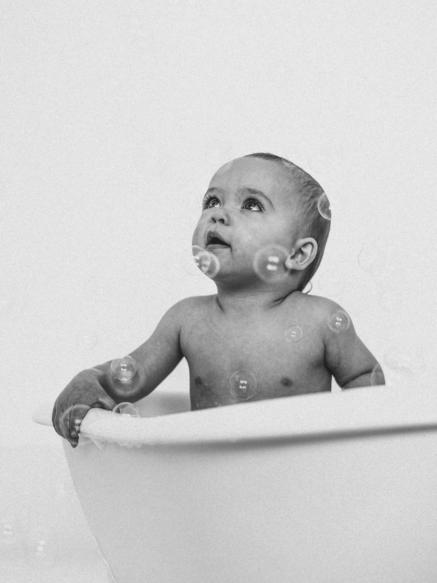 Black and white photo of a cute baby sitting in a bathtub surrounded by floating soap bubbles, looking up curiously.