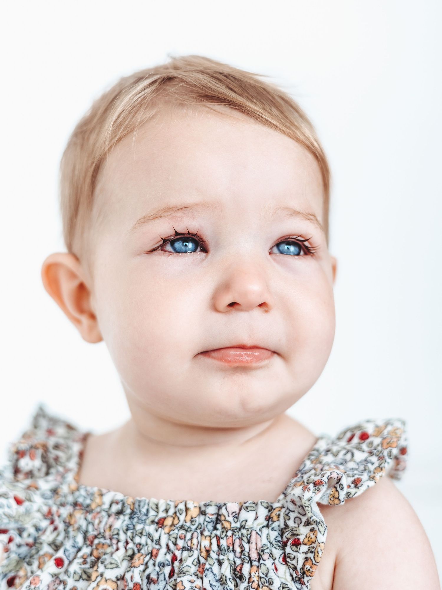 A baby wearing a floral dress gazes upward with bright blue eyes and a gentle smile.