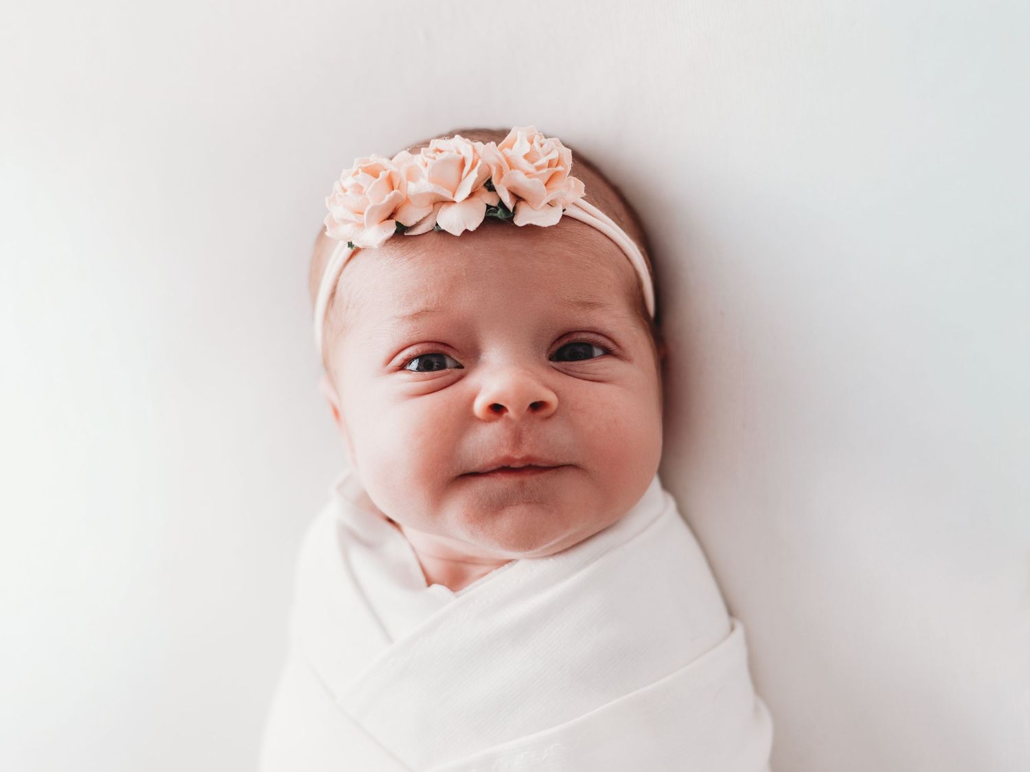 Newborn baby wearing white swaddle wrap and delicate pink floral headband while sleeping peacefully.