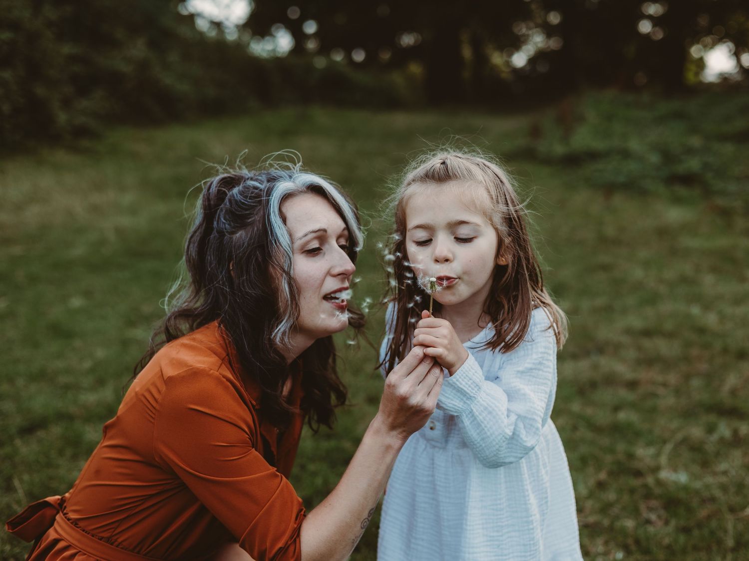 A parent and child share a tender moment outdoors while blowing on a dandelion together in a grassy field.