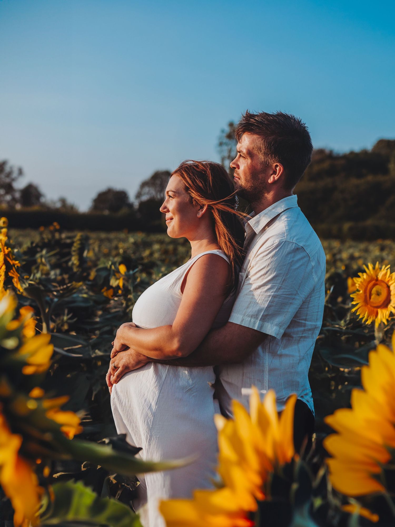 A couple embraces in a vibrant sunflower field during sunset, with the woman wearing a white dress and man in light shirt.