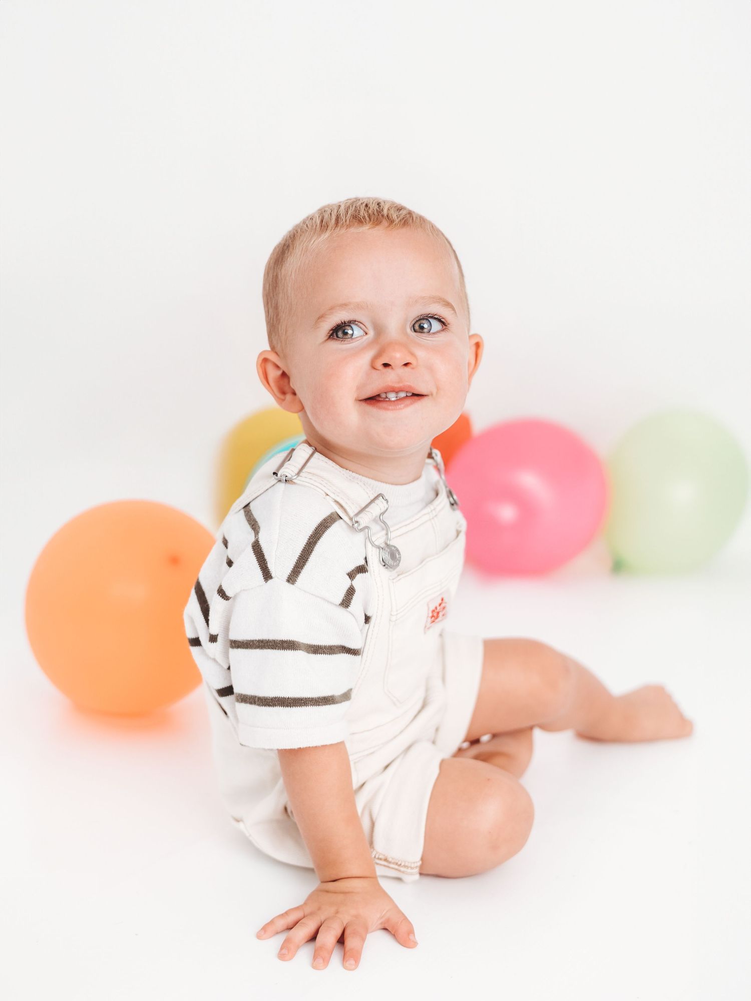 Happy baby sitting on white floor surrounded by colorful balloons, wearing striped onesie, smiling and looking up.