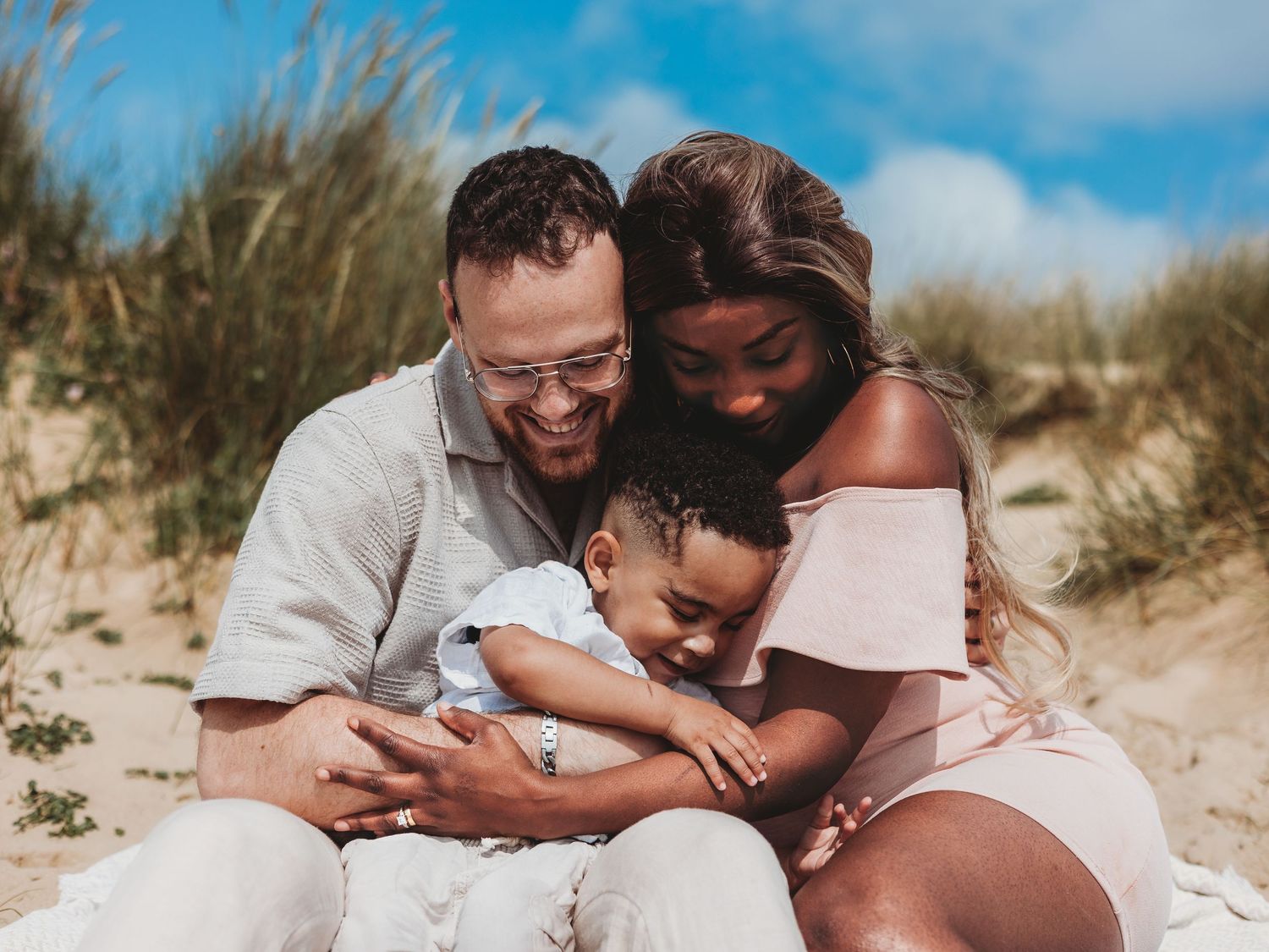 A happy interracial family with a baby sitting together on a sandy beach, smiling and embracing on a sunny day.
