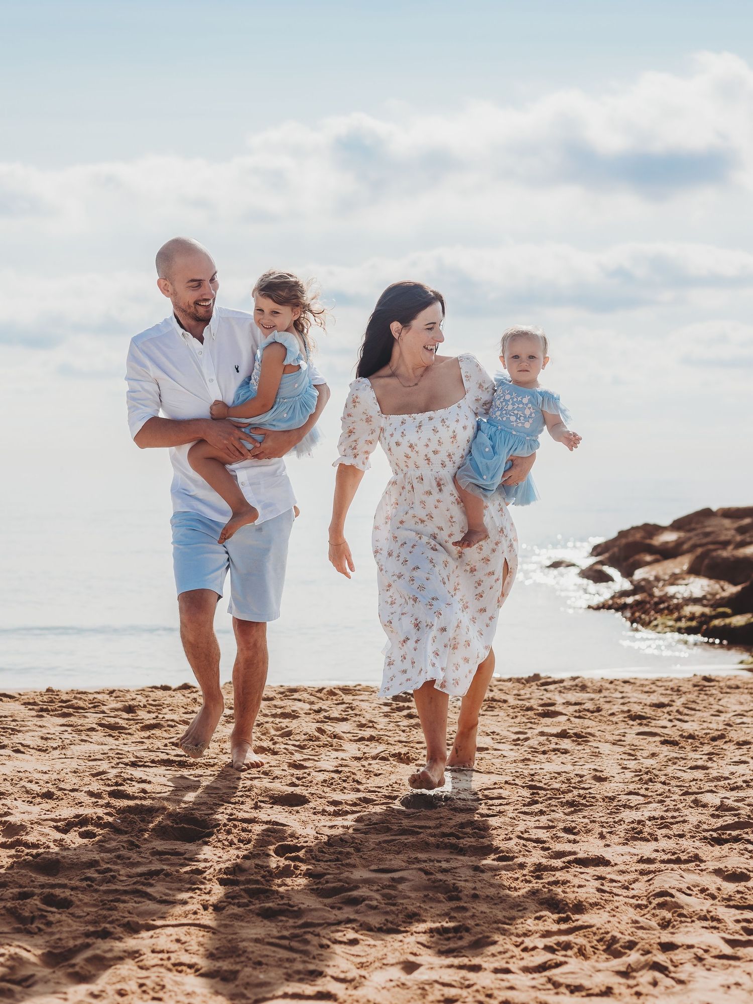 Happy family of four walking on a sandy beach, parents carrying young children, wearing light summer clothes on a sunny day.