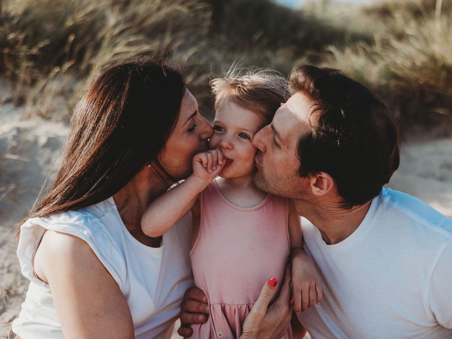 Parents kissing their smiling toddler girl on the cheeks at a sandy beach on a sunny day.