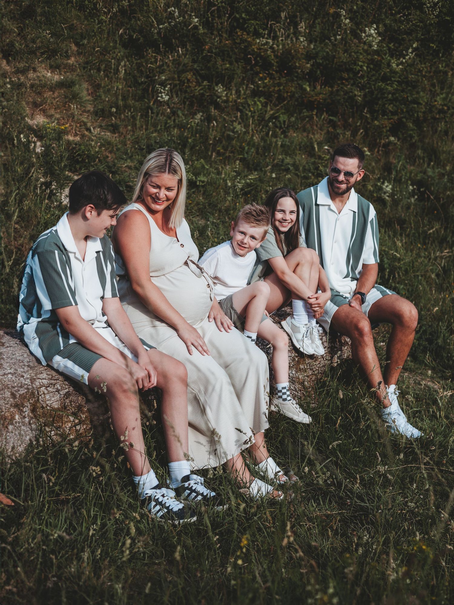 A happy family of five sits together on a rock in a grassy field, smiling and laughing in coordinated outfits.