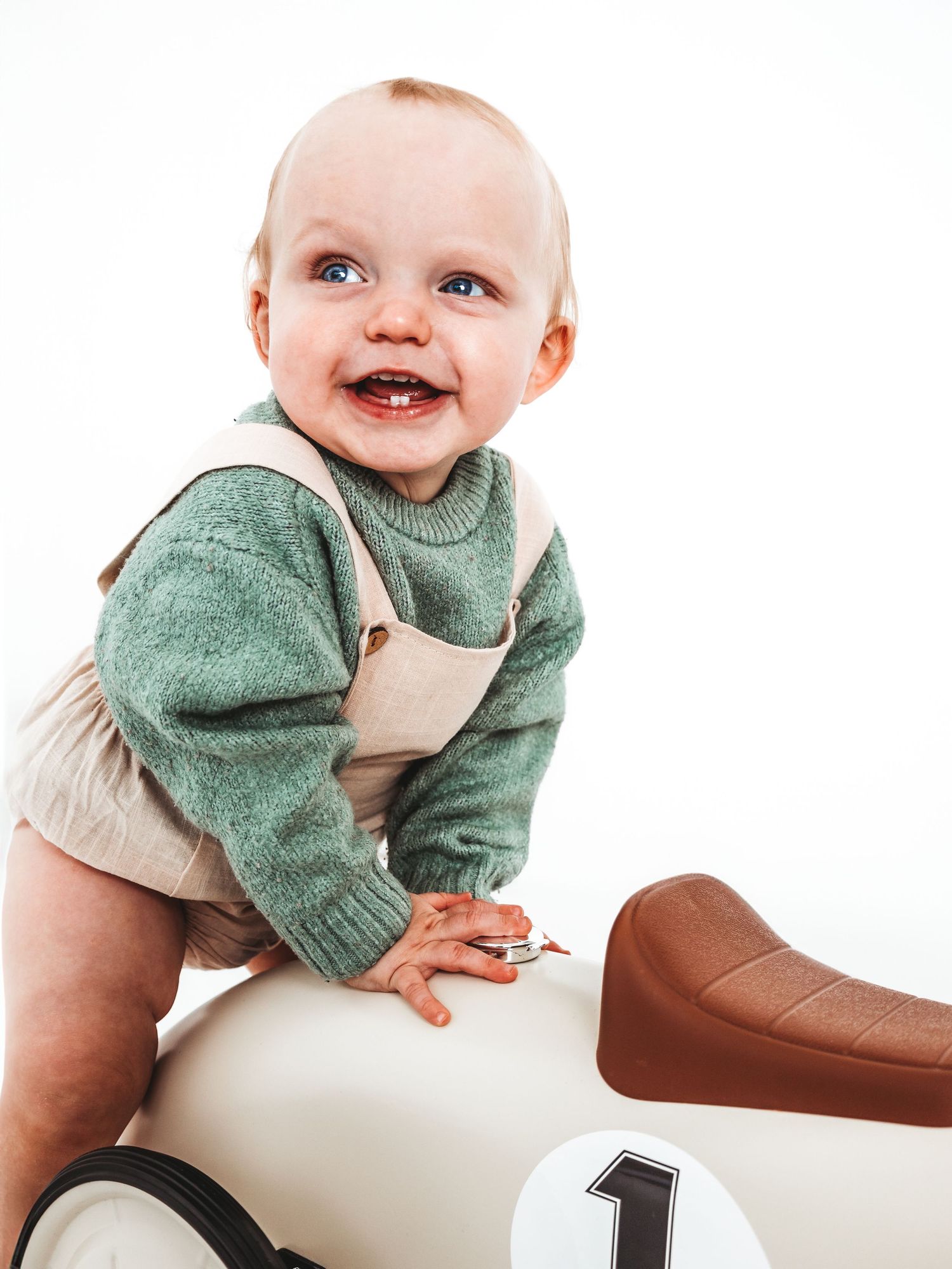 Happy baby in green sweater and overalls sits on a toy race car, smiling brightly against a white background.