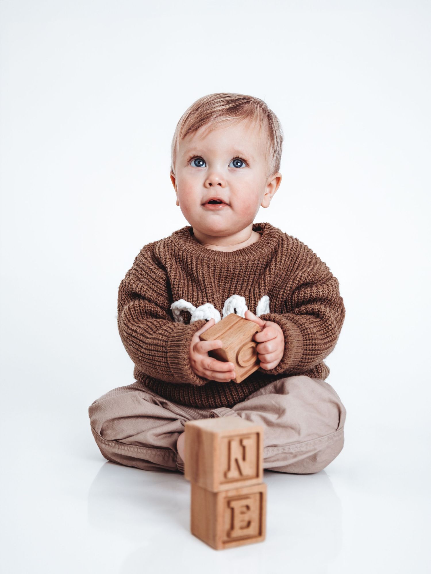 A toddler in a brown knit sweater sits cross-legged holding a wooden toy beside stacked alphabet blocks on white background.