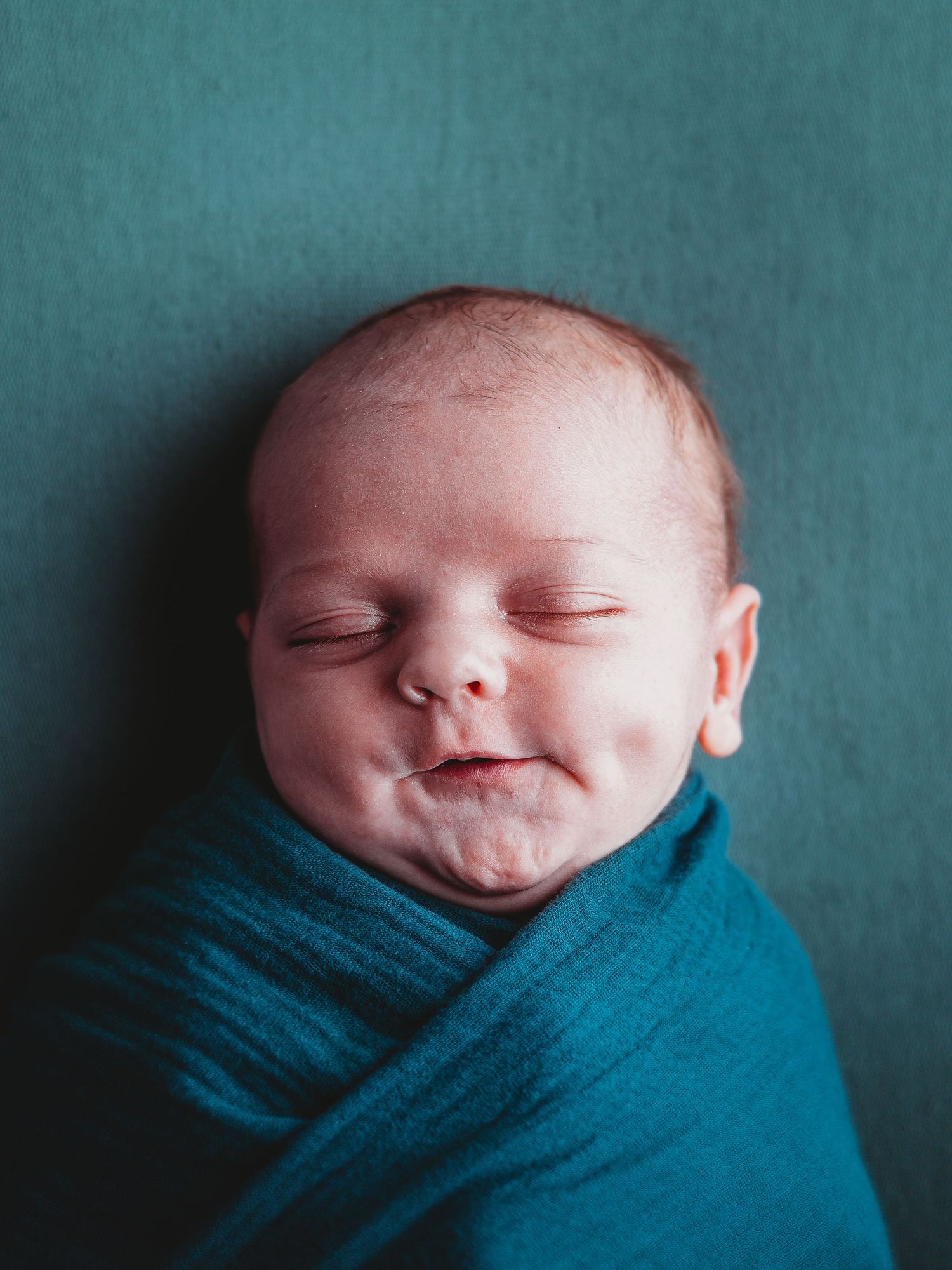 A sleeping newborn baby swaddled in a teal blue blanket against a teal colored backdrop.