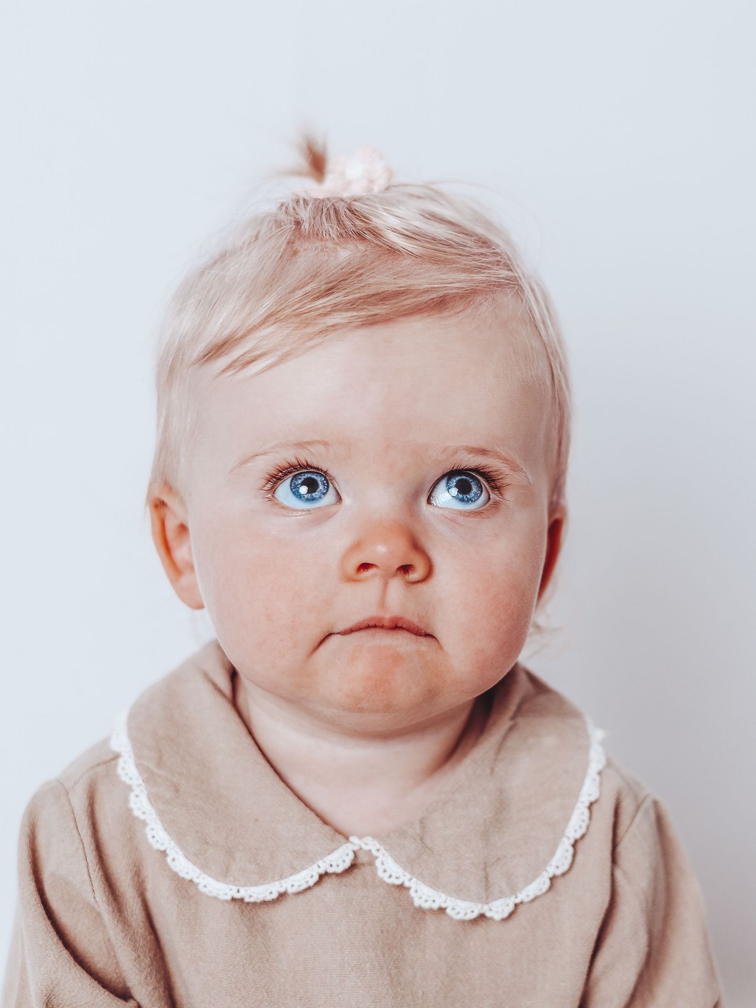 A baby wearing a beige collared top with white lace trim looks upward with bright blue eyes in a triptych portrait.