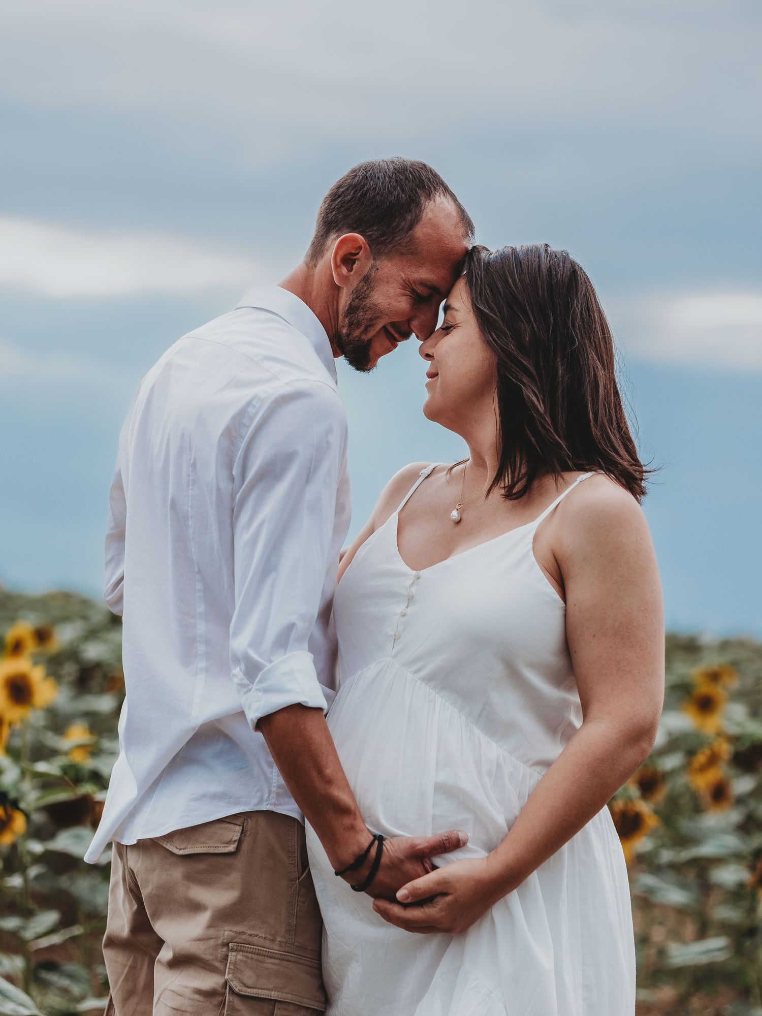 A romantic couple in white outfits poses together in a sunflower field on a summer day.