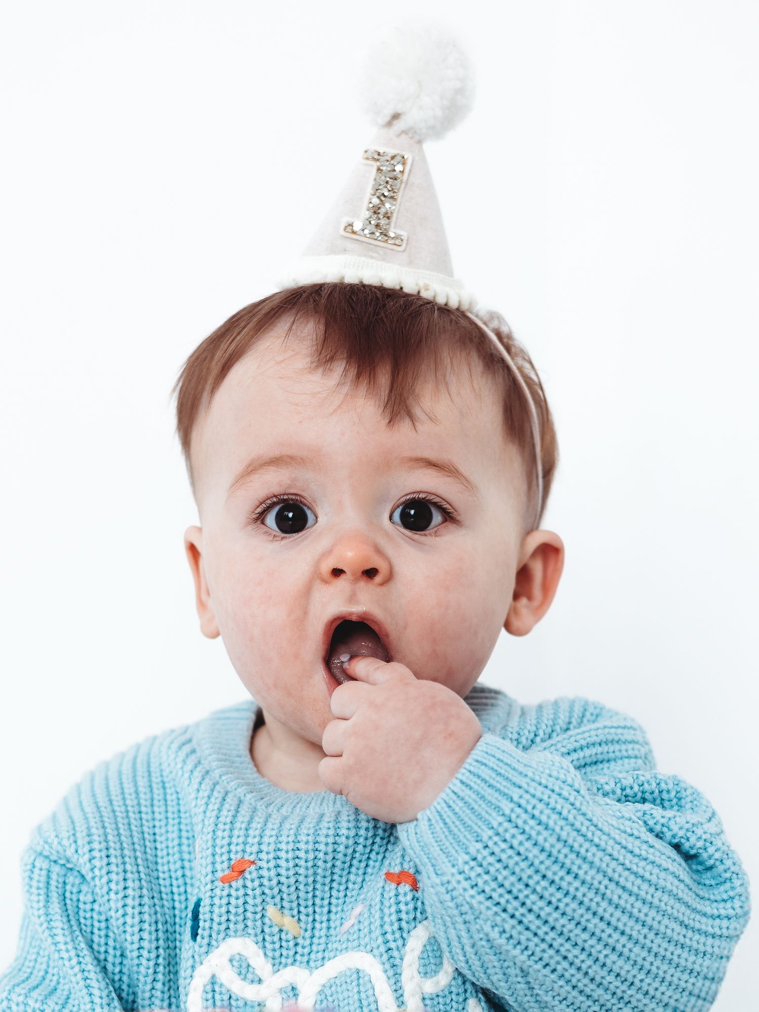 Cute baby wearing a white number one birthday hat and blue knit sweater, celebrating their first birthday.
