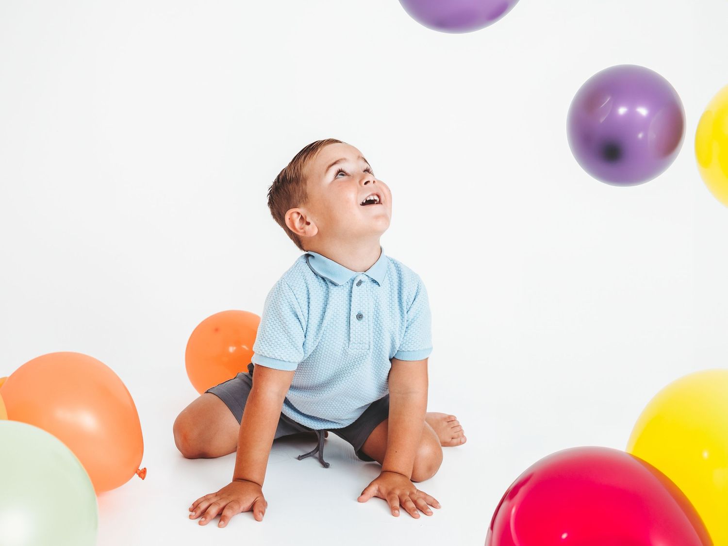 A joyful toddler boy sits on white floor surrounded by colorful balloons, looking up with delight.