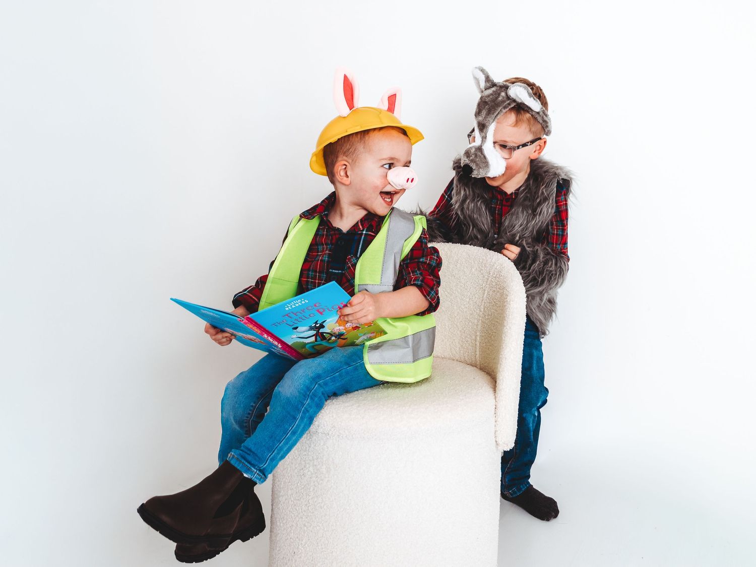 Two children in costumes, one as a construction worker reading a book, the other as a wolf, playing together on a white chair.