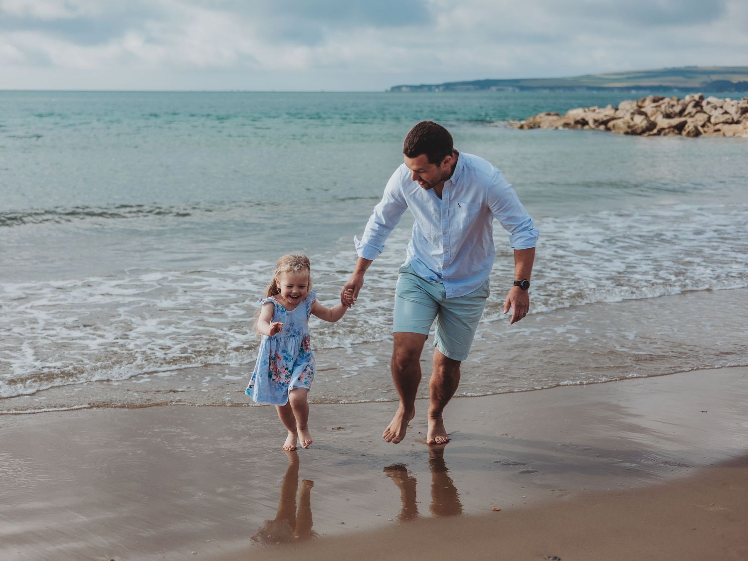 A father and young daughter hold hands while walking along a sunny sandy beach with gentle ocean waves.