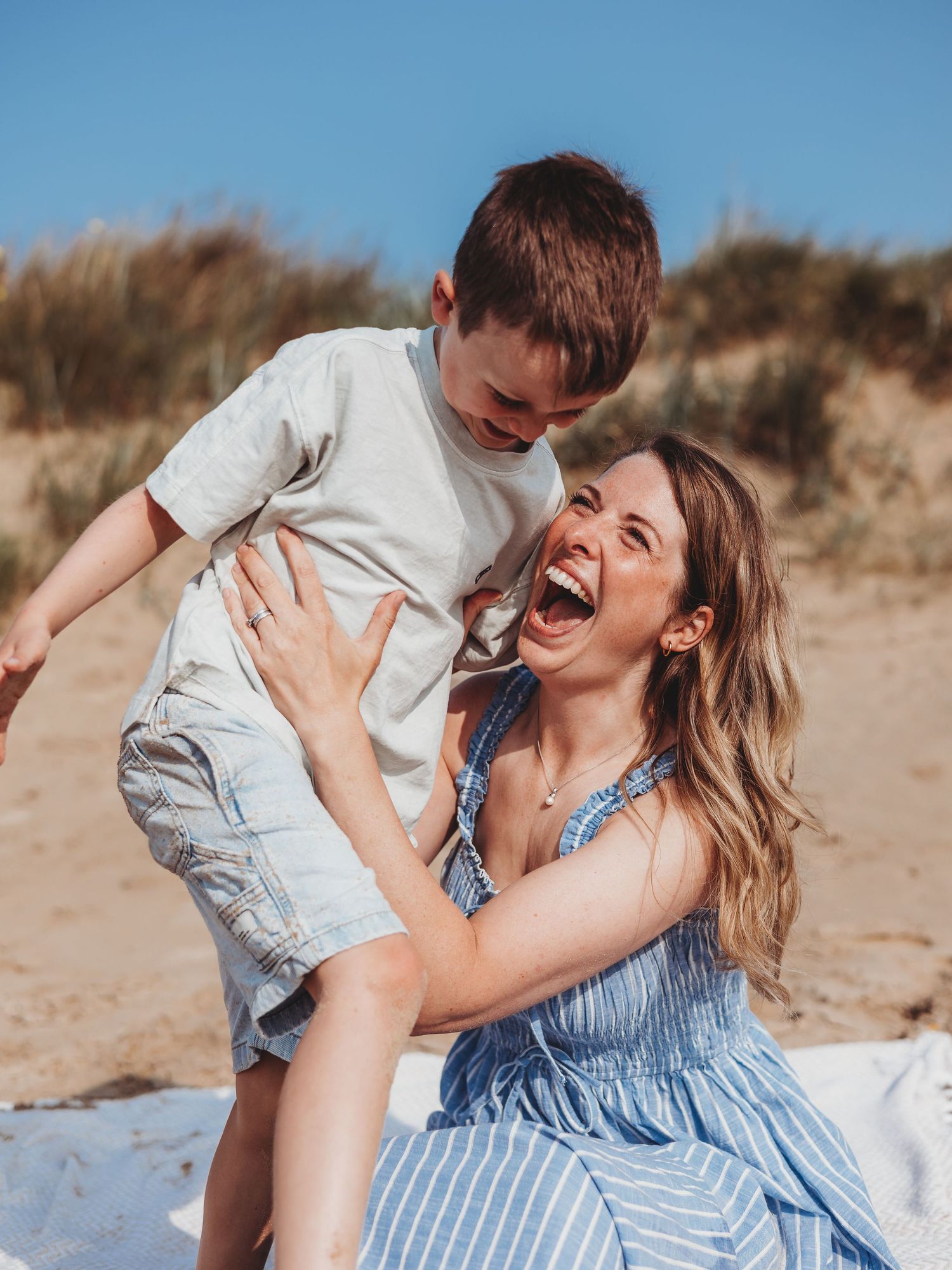 A joyful mother laughs while lifting her young son on a sunny beach, both sharing a playful moment together.
