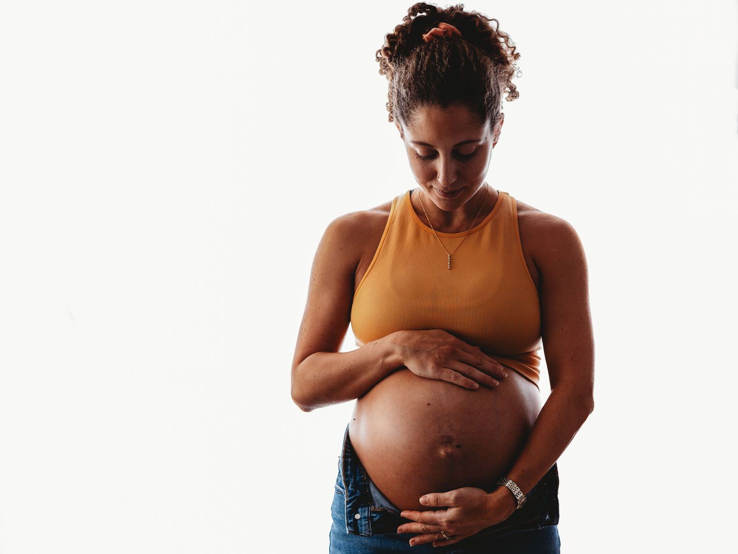Pregnant woman in brown tank top lovingly cradles her baby bump against a white background.