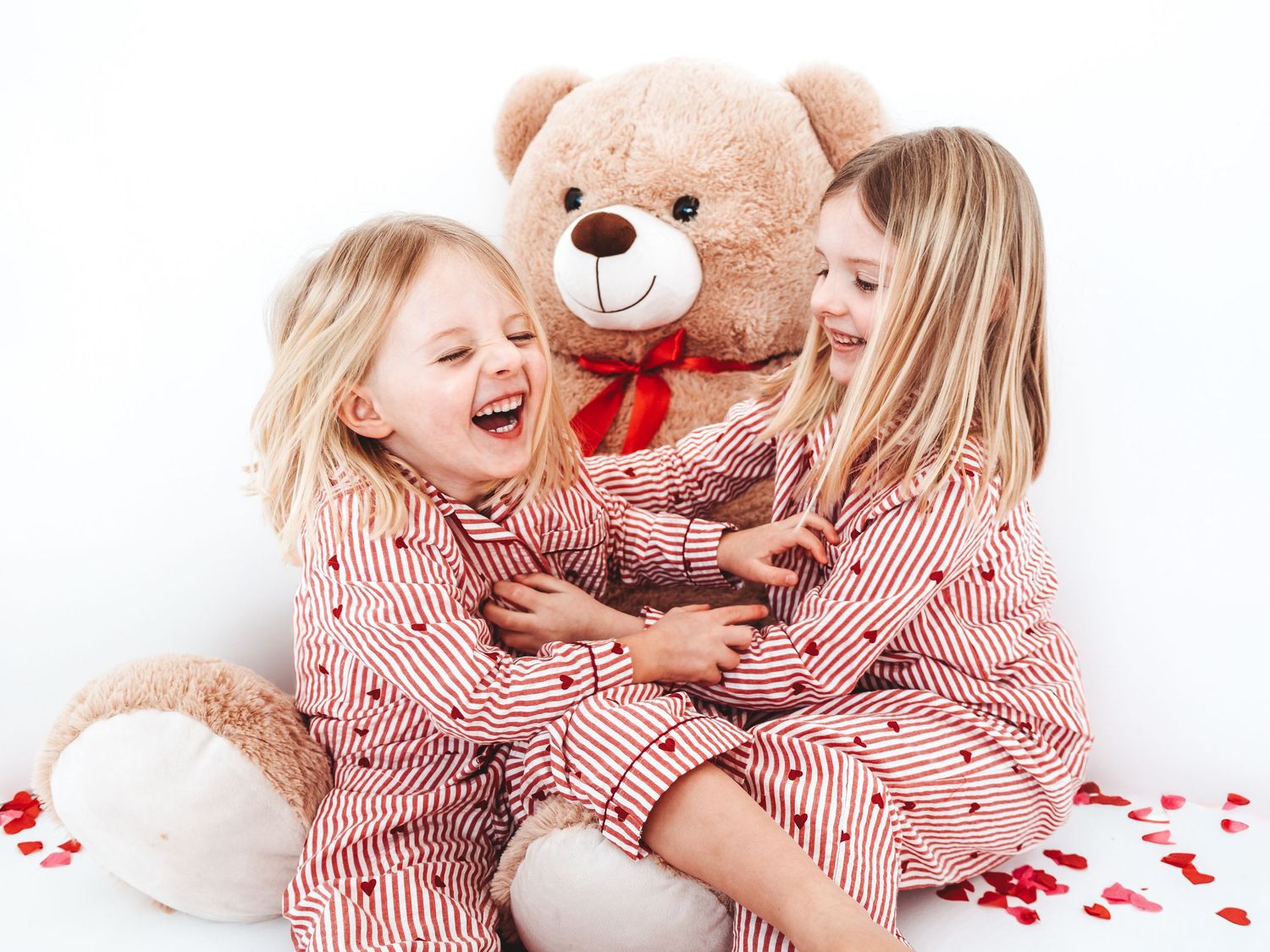 Two young blonde girls in matching striped pajamas laugh and hug a large teddy bear with a red bow.