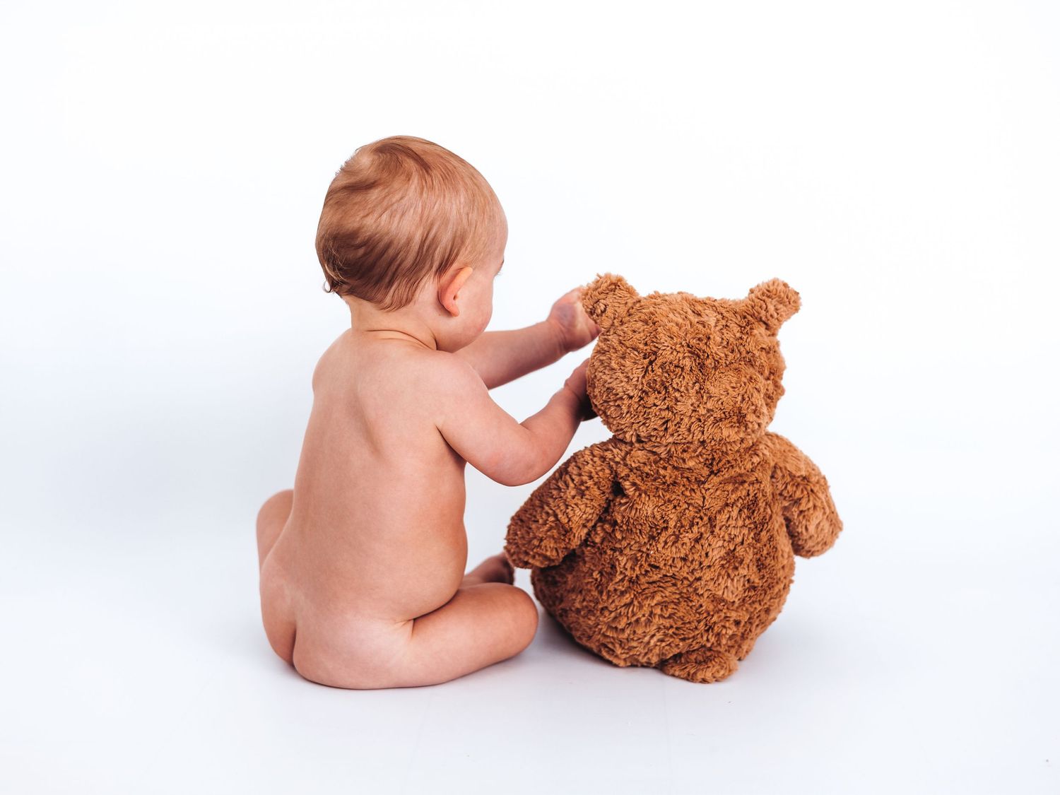Naked baby sitting with a brown teddy bear on a white background, viewed from behind.