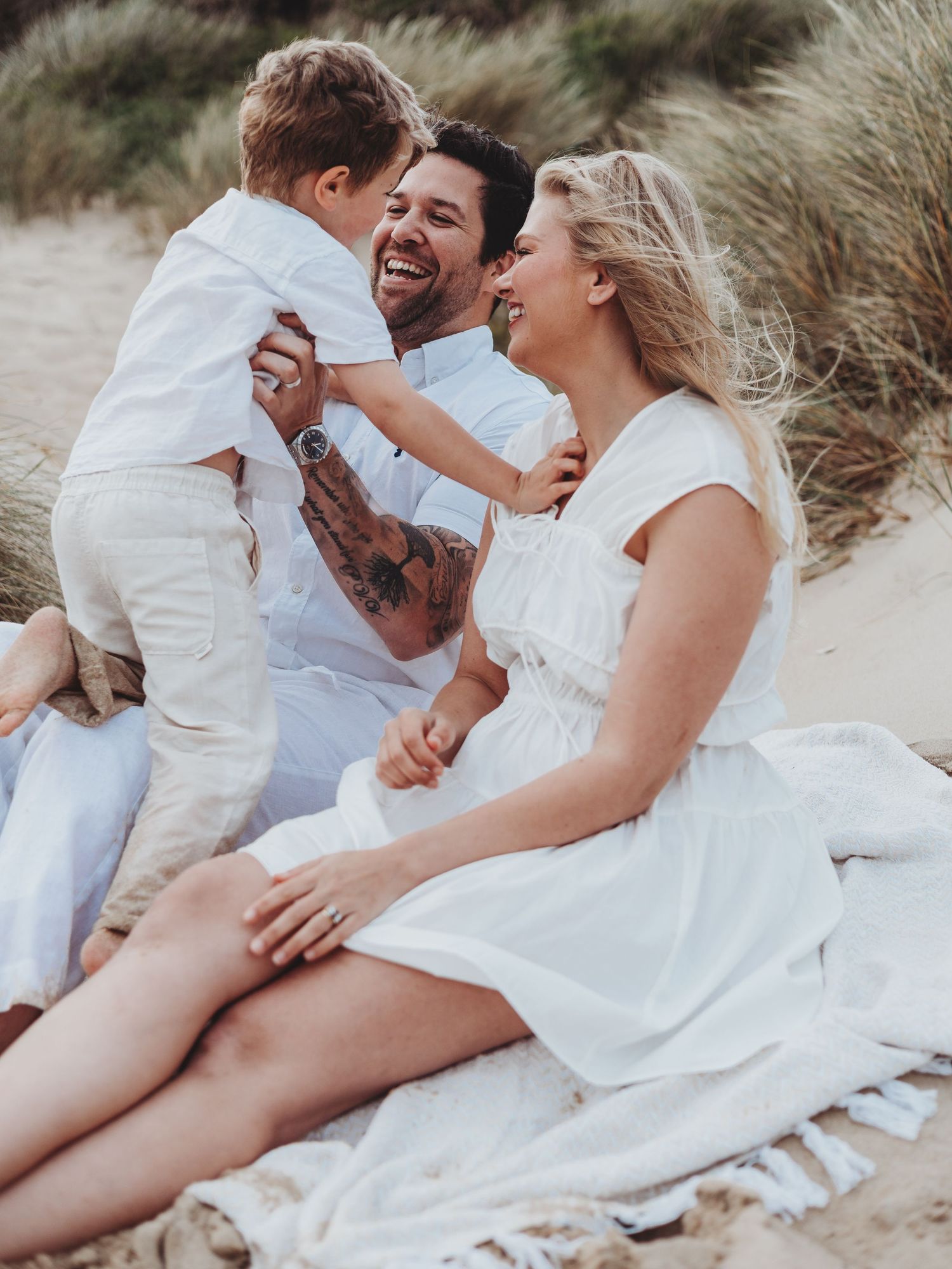 Happy family in white outfits laughing together on a sandy beach, parents playing with young son outdoors.